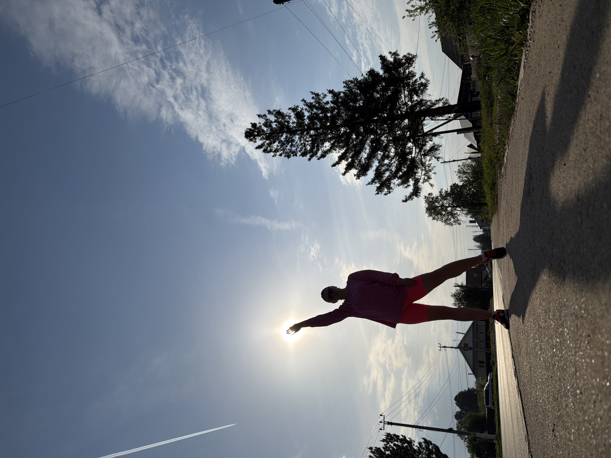 The photograph shows a person silhouetted against the sun in Chaikovsky, Russia. The person is standing on a paved road, facing towards the sun, with one arm extended as if reaching for it. The person is wearing athletic clothing consisting of a long-sleeved top and shorts. Overhead power lines are visible, along with trees and rooftops on either side of the road. There are clouds in the blue sky. The angle of the photo is tilted, with the road appearing to stretch from the bottom right corner to the upper left corner of the frame.