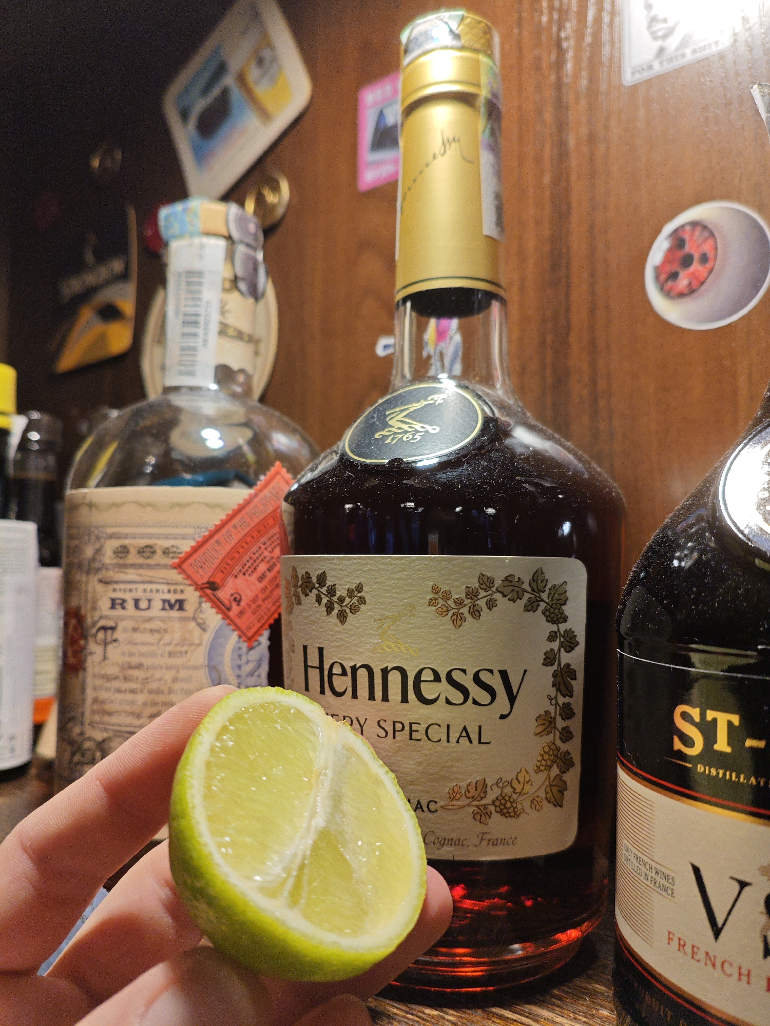 A close-up shot captures a hand holding a lime wedge in front of two bottles of liquor on a wooden shelf. The bottle on the left, a rum, is partially obscured. The bottle on the right is Hennessy VSOP. A third bottle is partially visible to the right of the Hennessy. The shelf appears to be in a bar or liquor cabinet. The image is taken indoors, with the lighting suggesting ambient light from overhead sources. The background is out of focus and appears to be a wooden surface with various stickers or labels. There is no discernible evidence indicating the location is in Kyiv, Ukraine.