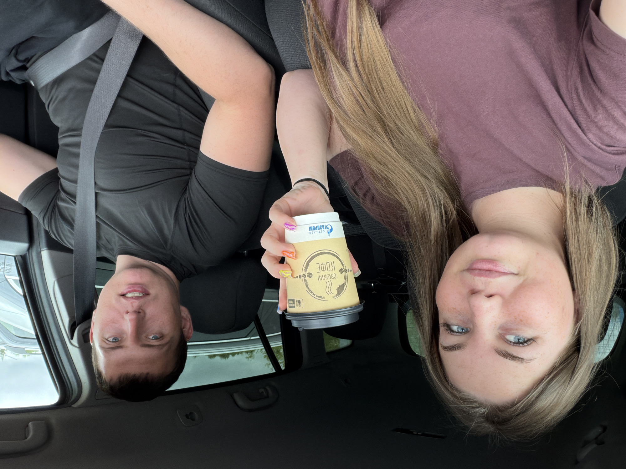 Inside a vehicle in Совхозный, Russia, a light-skinned female and a light-skinned male are seated. The female, positioned on the left, has long light brown hair, blue eyes, and visible freckles. She is wearing a dark mauve short-sleeved t-shirt and is holding a light brown paper coffee cup with a dark lid in her right hand. The cup features a "ГАЗПРОМ" logo and the Russian text "СВЕЖИЙ КОФЕ" (Fresh Coffee). Her fingernails are adorned with multi-colored polish. The male, seated on the right in the driver's seat, has short dark hair and blue eyes. He is wearing a black short-sleeved athletic shirt and a gray seatbelt across his chest and lap. Both individuals are looking towards the camera. The vehicle's interior appears dark, and through the windows, other vehicles and a cloudy sky are visible, indicating an outdoor setting.