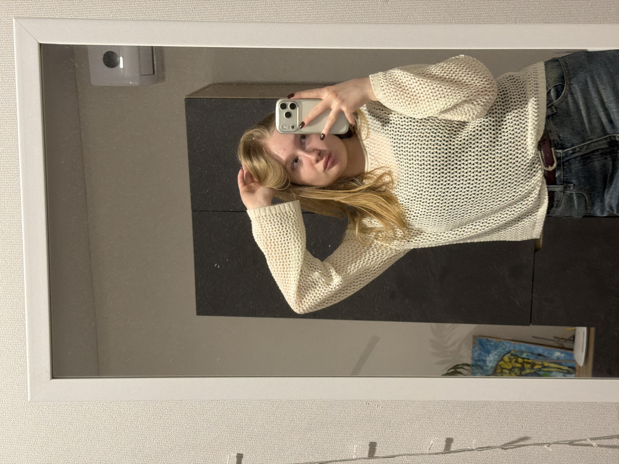 The image is a mirror selfie of a young woman wearing a white, long-sleeved, knitted sweater and blue jeans with a brown belt. She is holding a white smartphone in her right hand, taking the picture. Her left hand is near her head, touching her blonde hair.
Behind her, a dark grey cabinet is visible. In the upper left corner of the mirror's reflection, there's a white object, possibly a light fixture or device. In the bottom right corner of the mirror, there's what appears to be a painting or drawing with blue and yellow colors. The wall surrounding the mirror is a light beige color. The scene is indoors. The location in the city of Засечное, Russia cannot be determined from the image.