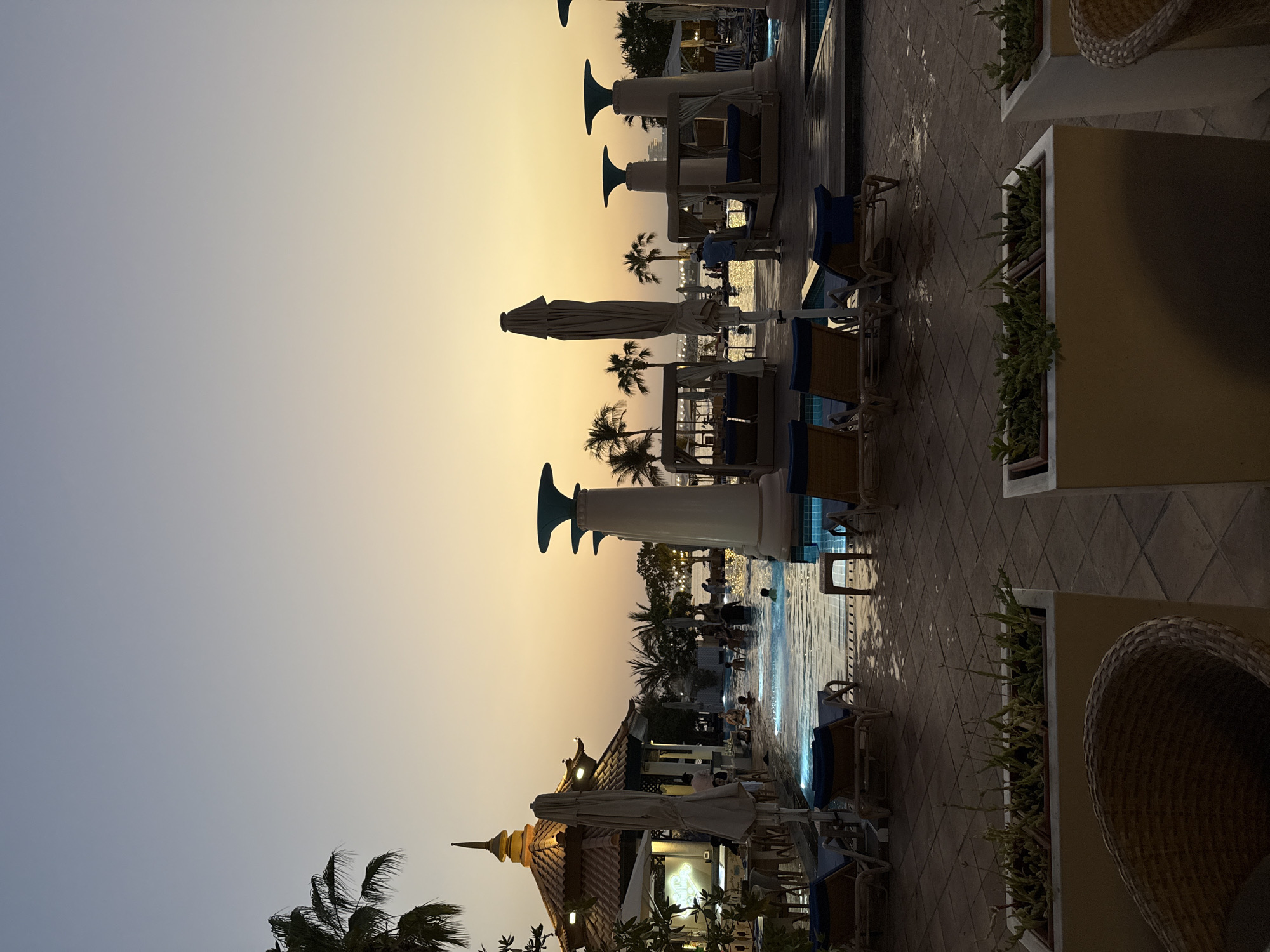 An outdoor swimming pool area at a resort in Dubai, United Arab Emirates, is depicted at dusk. The sky transitions from a light blue at the top to a soft yellow-orange near the horizon, indicating sunset. Palm trees are visible in the background, along with a building with a distinctive tiled roof, resembling traditional Asian architecture, possibly a restaurant or bar. Several lounge chairs are arranged around the pool, some with blue cushions. In the pool, several people are swimming and enjoying the water. Two cabana-style seating areas with white curtains are positioned near the pool. A man in a blue shirt and beige pants is standing near one of the cabanas, looking towards the pool. The ground around the pool is tiled and appears wet in places. Planter boxes with green foliage are in the foreground. The overall atmosphere is tranquil and relaxed, characteristic of a resort setting during the evening.