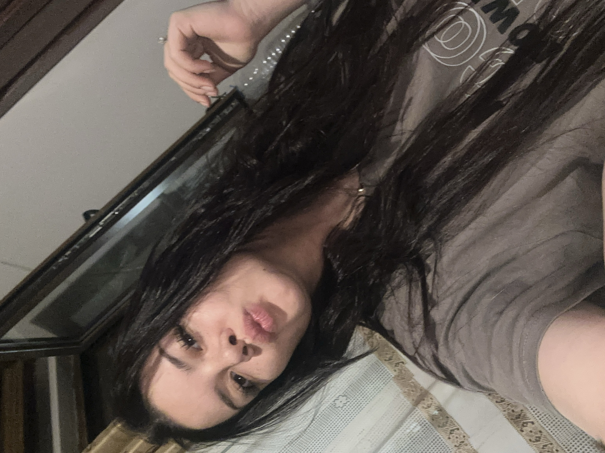 An indoor selfie taken in Chubra, Bulgaria, depicts a person with long dark hair and fair skin, captured through a mirror. The image is oriented sideways, suggesting the person is reclining or lying down on a surface covered by a patterned fabric, possibly a bedspread with stripes and decorative motifs.
The subject is wearing a dark gray t-shirt that features white graphic elements and partial letters, including "MO" and circular designs, on the upper chest. A hand with a ring on one finger is visible near the upper left, seemingly interacting with the mirror's edge or hair. The person's face is central, with a playful or casual pout evident on the lips.
The background, visible through the mirror, consists of a plain light-colored wall or ceiling and a dark wooden frame, possibly part of a window or furniture. The lighting is soft and ambient, indicative of an indoor setting, likely evening or under artificial light. There are no clear external weather cues or specific time of day discernible beyond the indoor context. No other distinct objects are prominently featured.