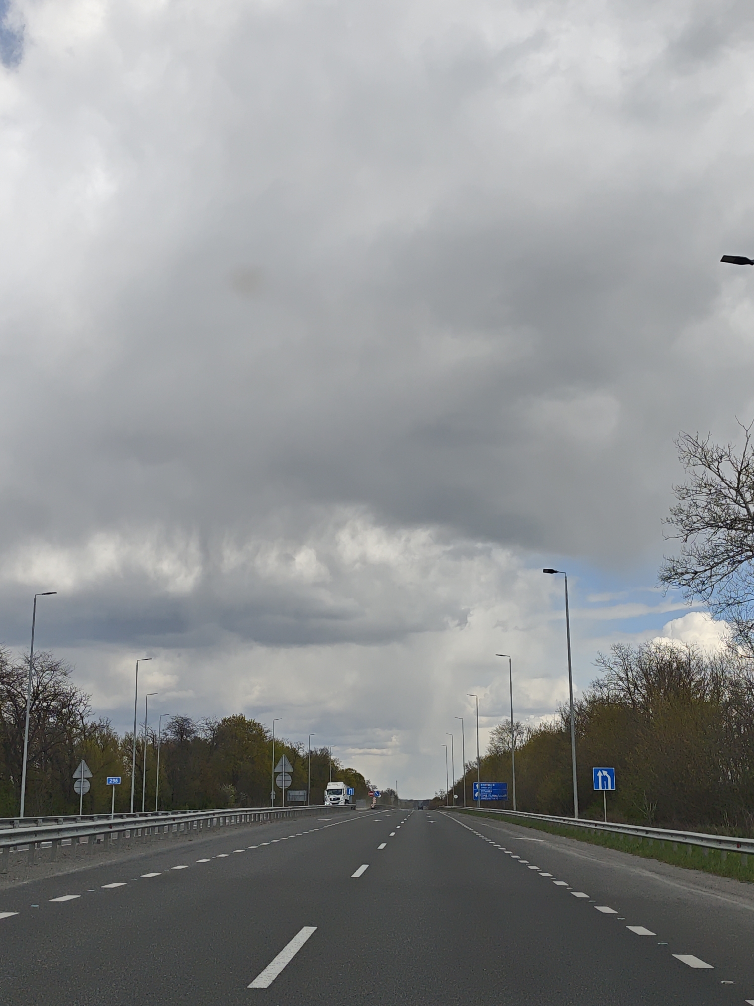 This image presents a view from a vehicle traveling on a multi-lane highway in Holoskiv, Ukraine. The asphalt road stretches into the distance, appearing largely empty except for a white semi-trailer truck visible in the right lane in the background, moving away from the viewer.
The sky is a dominant feature, heavily overcast with dark grey clouds that suggest impending rain or a general somber weather condition. However, patches of blue sky are visible towards the horizon and on the far right, indicating variable weather. Tall, dark grey streetlights line both sides of the road, extending into the perspective.
The sides of the highway are flanked by green vegetation, including lush trees and dense bushes. Some trees appear to have bare branches, suggesting early spring or late autumn. A metal guardrail runs along the right side of the road.
Several road signs are visible:
- On the left, a blue rectangular sign displays the number "296" in white text.
- On the right, a blue square sign with a white U-turn arrow symbol is clearly displayed.
- Further in the distance, other blue directional signs are present on both sides, but their text is not legible.
The road itself features clear white dashed lines separating lanes and solid white lines marking the shoulders, with reflective markers embedded. The time of day is daytime, likely mid-morning or afternoon, given the natural light despite the heavy cloud cover. The scene conveys a sense of travel on a relatively quiet road under a dramatic, cloudy sky.