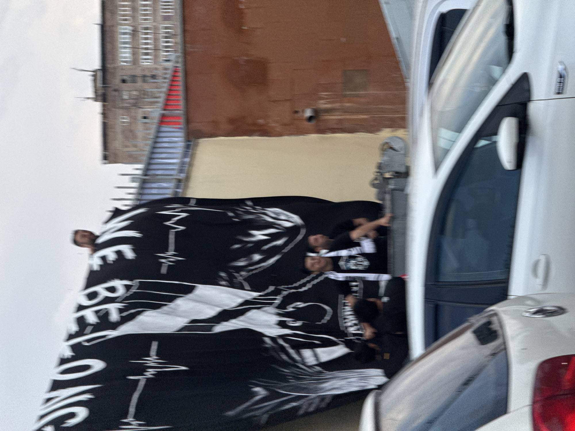 A banner with white lettering on a black background, displaying a heartbeat line and a graphic of a face, is held aloft by several people standing on a roof. The banner is partially obscuring a brick building. In the foreground, two white cars are parked, one of which partially obscures the view of the other. The scene appears to be set in a city environment, with the architecture suggesting a location in Abovyan, Armenia. The sky is overcast.