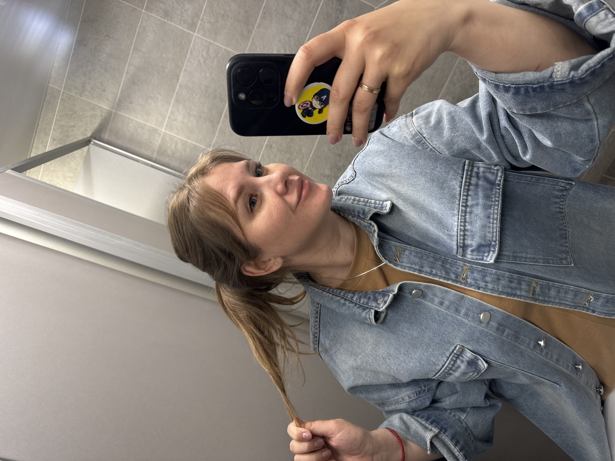 A casual mirror selfie captures a young adult woman in an indoor setting, likely a public restroom or changing room, based on the tiled walls and reflective surfaces. The woman has light brown hair, pulled back into a low ponytail, and light-colored eyes. She wears a light blue denim jacket over a brown or tan t-shirt and a thin silver necklace. On her left ring finger, she wears a simple gold ring, and on her right wrist, a red string bracelet is visible.
She holds a black smartphone, likely an iPhone with multiple camera lenses, in her right hand to take the photo. A yellow PopSocket featuring a cartoon depiction of Captain America holding his shield is attached to the back of the phone. Her left hand is casually resting on a strand of her ponytail. The woman's expression is neutral and calm, with a slight, subtle smile, as she looks towards the phone/mirror.
The background shows light grey rectangular tiled walls. A white or light grey partition with a silver metallic trim runs across the lower portion of the image behind her, suggesting a stall or changing area. The image is tilted, indicating the angle at which the selfie was taken. No explicit text is visible beyond the recognizable character on the phone accessory. The context provided indicates the location is Perm, Russia.