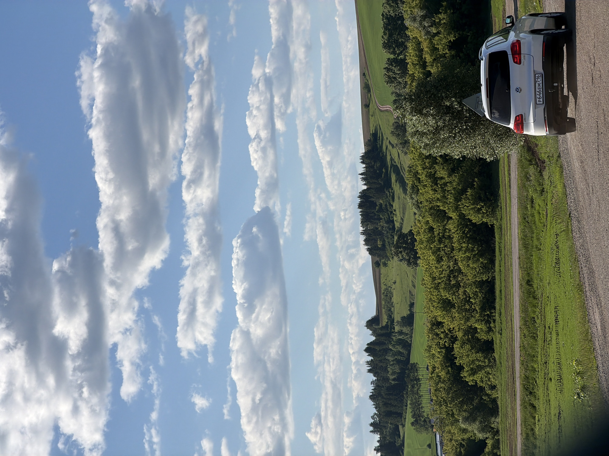 The image captures a rural landscape on a clear, partly cloudy day. The location is Unknown, Unknown. The sky occupies the upper half of the frame, displaying a vibrant blue punctuated by numerous white and grey cumulus clouds of varying sizes, arranged horizontally and scattered. The lower half of the image features undulating green hills and fields, some appearing as cultivated agricultural land, others covered in lush grass. Patches of dense deciduous and coniferous trees are visible across the hills, forming distinct forest lines.
In the foreground, a light-colored gravel road extends from the bottom right towards the center, where a white Volkswagen Tiguan SUV is parked on the right side, facing away from the viewer. The vehicle has a Russian license plate, which reads "P646EM 716". A triangular road sign, partially obscured by the vehicle and a nearby tree, is situated to the left of the car. Behind the car and road, a thick line of green trees extends across the middle ground. Further in the background, a narrow, winding dirt path or road is discernible on a distant green hillside, climbing towards the horizon. The sunlight suggests it is daytime, casting clear illumination on the scene. No people are visible.