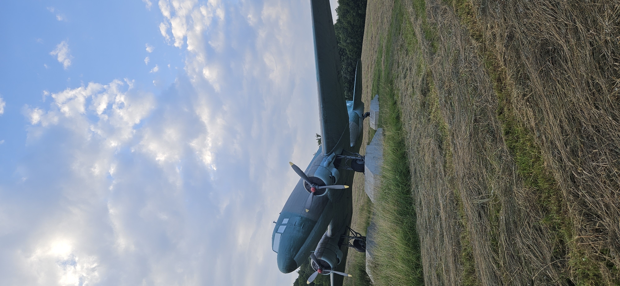 An old, teal-colored propeller airplane is parked on a grassy field in Vyšný Komárnik, Slovakia. The airplane is supported by concrete blocks and sits on landing gear. The sky above is blue with scattered white clouds, and the sun is partially visible. The field is covered in dry, matted grass, with some green grass peeking through. Trees are visible in the background. The scene appears to be a static display of the aircraft.