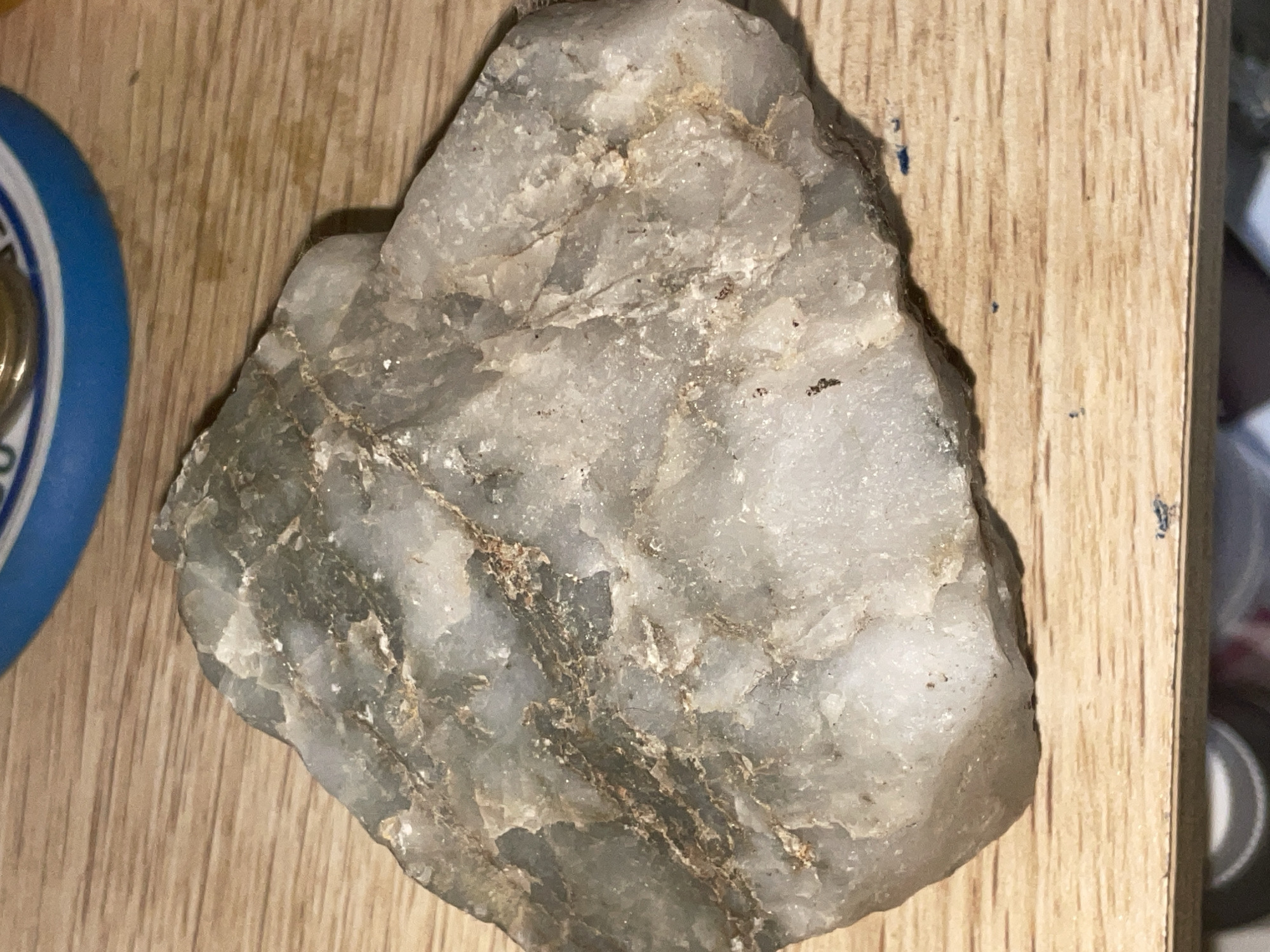 The image shows a rough, gray and white rock with brown and green veins. It is resting on a wooden surface. In the upper right corner, a blue and silver object is partially visible, possibly a lid or container. There are also some small dark specks on the wooden surface, and what appears to be a tiny dark blue smudge. The image is a close-up of the rock and does not provide any information about its location in Davao City, Philippines, or any other context.
