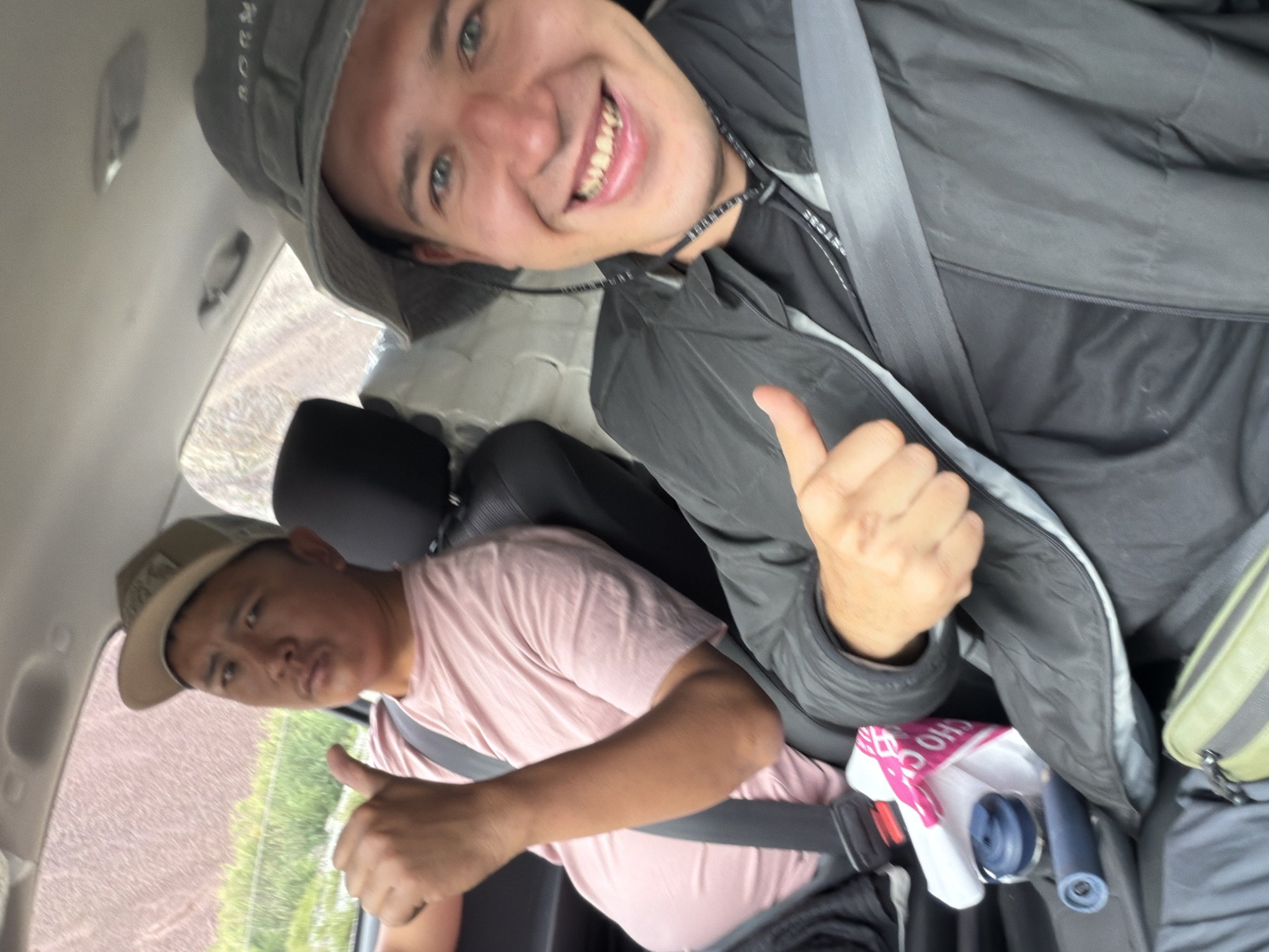 Two men are in the front seats of a car. The man in the driver's seat, wearing a pink t-shirt and a tan baseball cap, is giving a thumbs-up. The passenger, wearing a black jacket and a gray bucket hat, is also giving a thumbs-up and smiling. The car appears to be driving on a dirt road, with a brown, rocky hillside visible outside the window. There are bags and a water bottle on the floor of the car. The image is taken from a low angle, looking up at the two men.