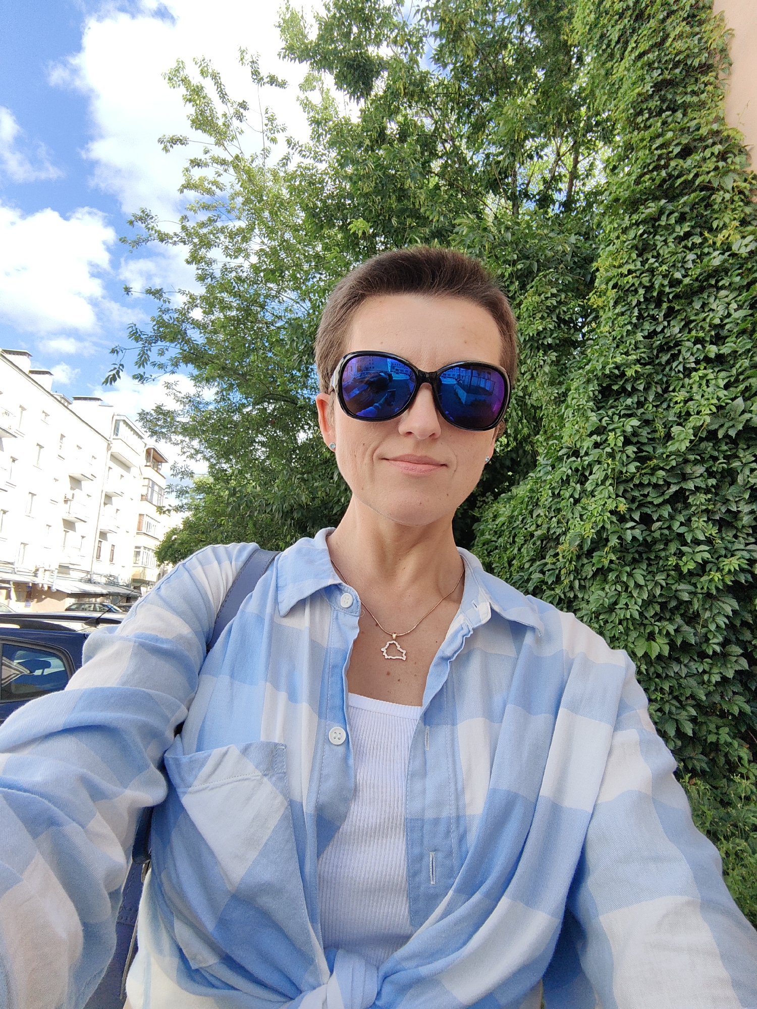 A woman with short brown hair and sunglasses is taking a selfie outdoors. She is wearing a blue and white checkered unbuttoned shirt over a white tank top and a thin gold chain with a pendant. She is standing on a city street with a building to her left and a wall covered in green ivy to her right. The sky above is blue with white clouds. The overall environment suggests a warm, sunny day in an urban setting. The city of Minsk, Belarus, is mentioned as the location.