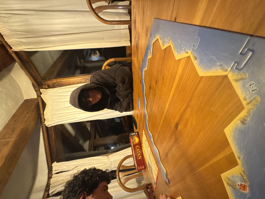 Two people are at a wooden table playing Catan. One person is wearing a dark hoodie and sitting near a window with white curtains. The game board is partially assembled with a blue edge. The Catan box is visible next to the board. Part of the second person's face and hair are visible. The room appears to be lit with warm light.