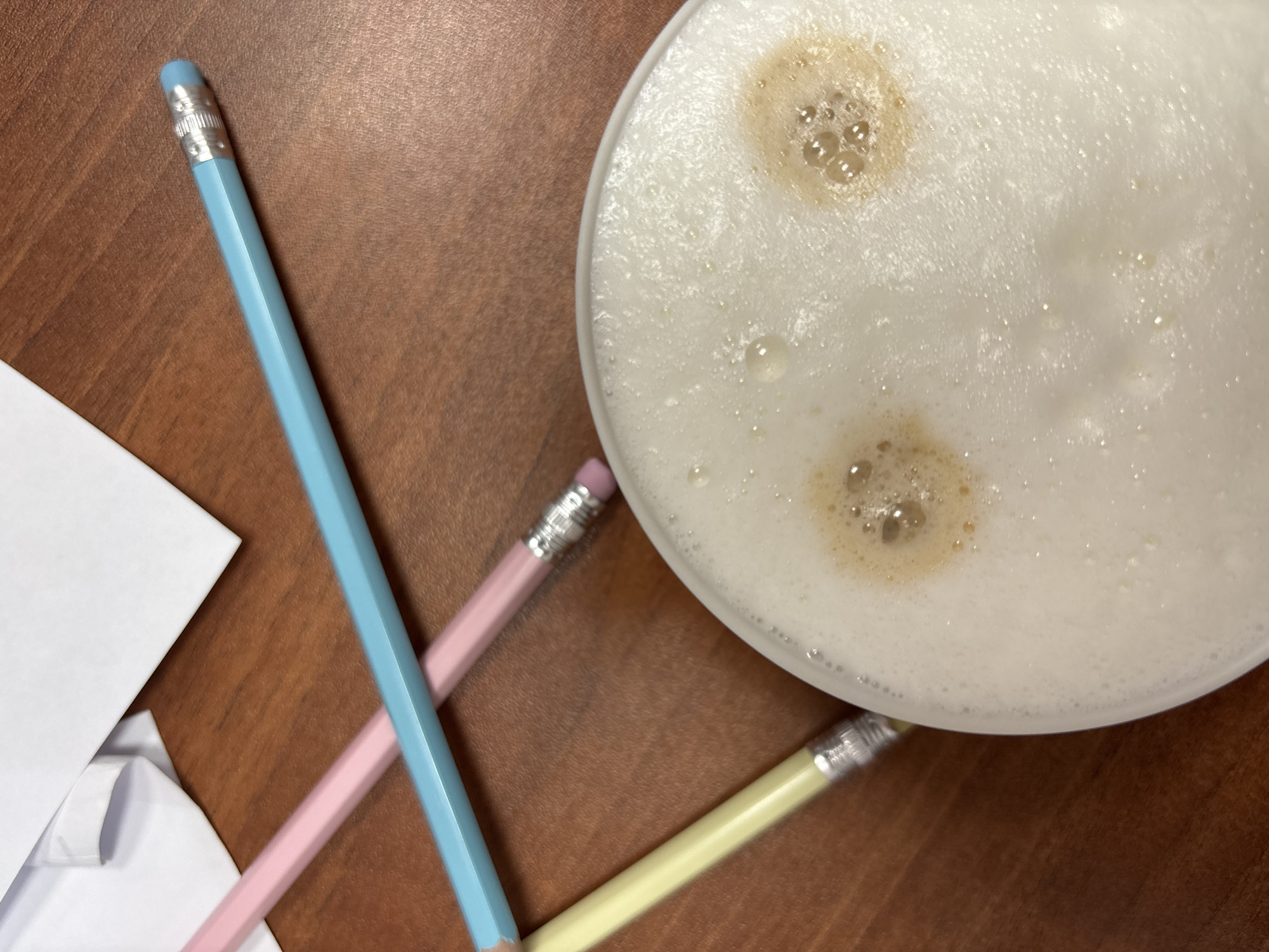 The image shows a close-up of a wooden table in an indoor setting. A partially visible white cup filled with a foamy beverage (likely coffee or a latte) dominates the right side of the frame. Three colored pencils—light blue, pink, and yellow—are arranged diagonally across the surface, their ends near the cup. To the left, a stack of white paper is partially visible. There is no evidence indicating the location is Chaikovsky, Russia. There are no people, actions, or interactions visible.