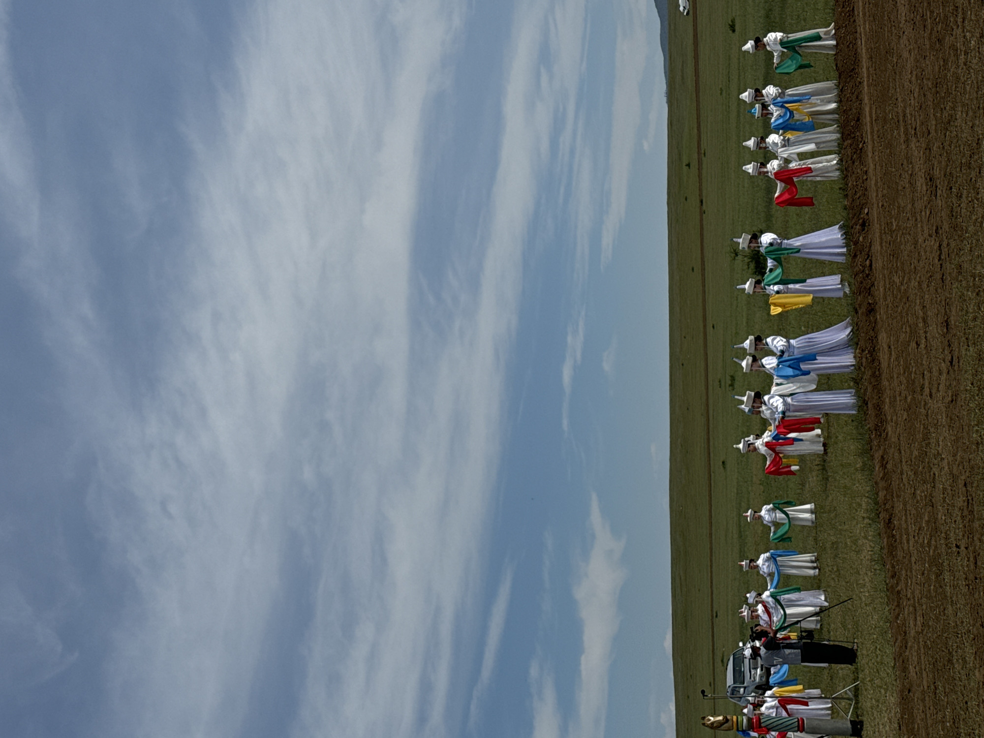 The image depicts an outdoor event in Orongoi, Russia, featuring a group of individuals dressed in traditional white garments with distinctive hats. They are holding colorful sashes in various hues, including blue, green, yellow, and red, and appear to be performing a coordinated dance or ritual in a grassy field. Some of the individuals are holding their sashes in flowing arcs. In the background, a silver SUV is parked. A cameraman with a video camera on a tripod is positioned to the left of the group, actively filming the event. The scene is set against a backdrop of rolling green hills under a cloudy blue sky. A partially tilled field is visible in the foreground.