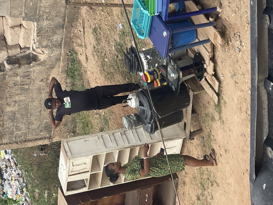 A man in black clothing poses next to a large, black, cylindrical object. A woman wearing a green dress and sandals stands near a white shelving unit, looking at her phone. The background includes other objects like colored buckets, stacked furniture, and a pole running through the scene. A pile of waste is visible in the distance.