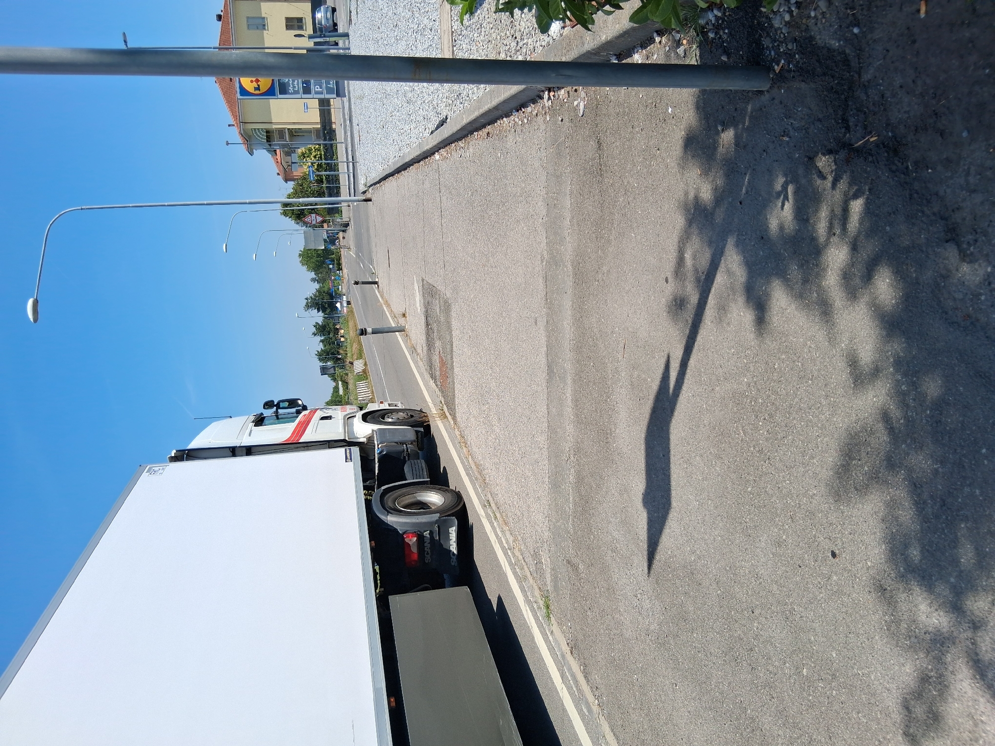 A semi-trailer truck, branded "SCANIA," is parked on the side of a road with its trailer extending towards the left of the frame. The truck is white with a red stripe and the word "SCANIA" is visible on the mud flaps. The truck is on a paved surface, adjacent to a sidewalk. The sidewalk is concrete and runs alongside a gravel parking lot. In the background, there is a LIDL supermarket with its distinctive blue and yellow logo. Several streetlights are visible along the road, which appears to be a main thoroughfare. The sky is clear and blue, indicating a sunny day. The scene is devoid of people. The location is the Via Gandini in Colorno, Italy, in front of the LIDL supermarket.