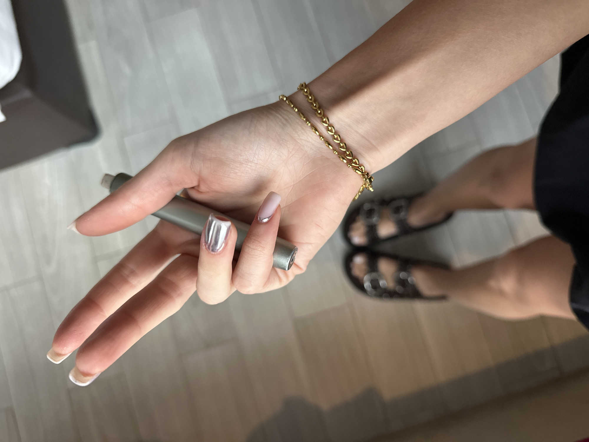 A close-up, high-angle shot depicts a person holding a silver-colored cylindrical object. The person's left hand and lower right leg are visible. The person has long fingernails with a light pink base coat. The nail on the ring finger is covered in silver nail polish. A delicate gold chain bracelet adorns the wrist. The person is wearing dark sandals. The setting appears to be indoors on a light-colored wooden floor. A piece of furniture is partially visible in the upper left corner. There is no indication of the city or country.