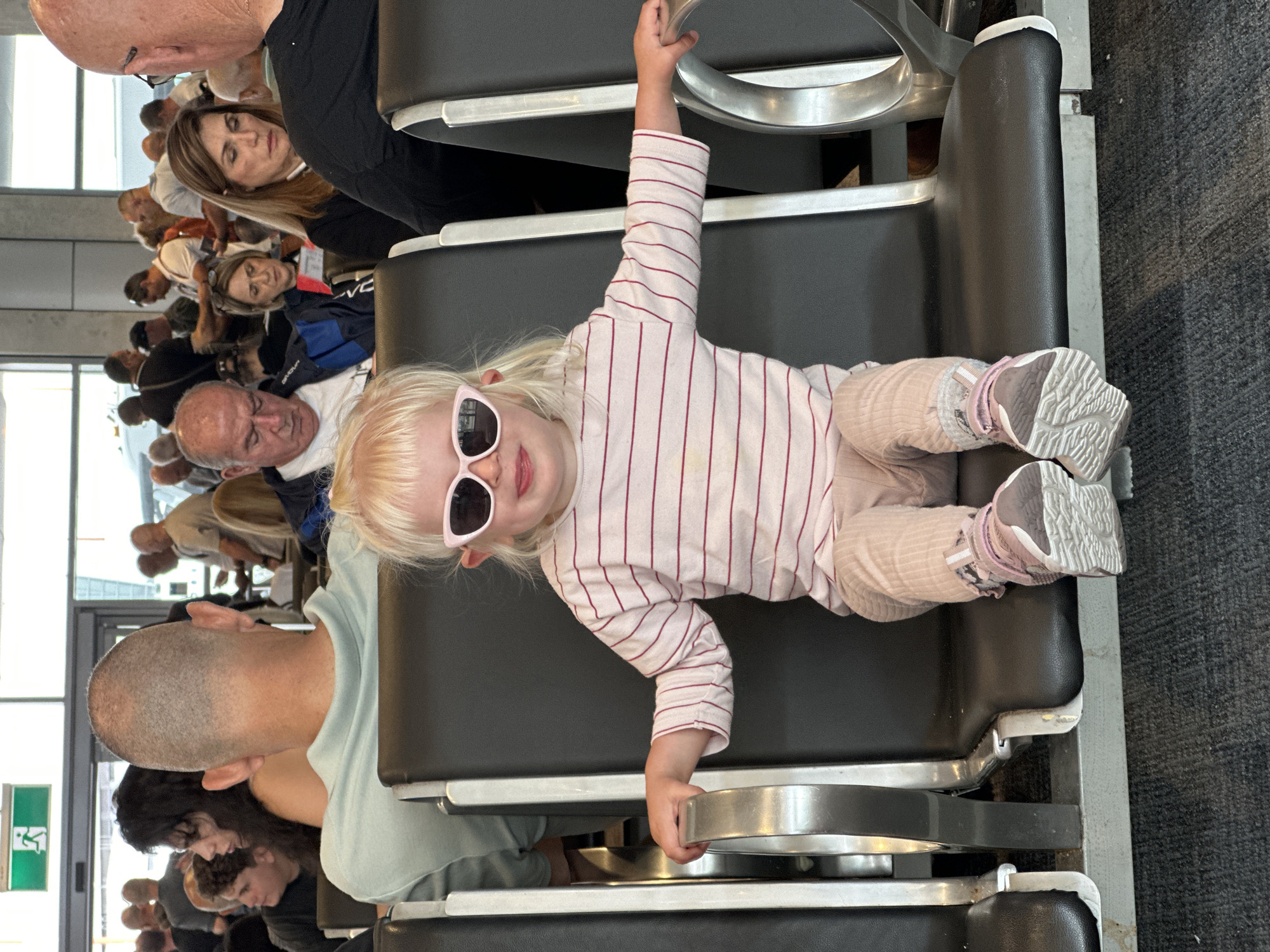 A young child with blonde hair and pink cat-eye sunglasses is sitting in an airport waiting area, holding onto a metal seat divider. The child is wearing a striped long-sleeved shirt and light pink pants, with pink and grey sneakers. Several other people are visible in the background, seated and standing, some facing away from the camera. The environment appears to be an indoor public space with dark grey seats and carpeted floor. An exit sign with a green background and a white figure is visible in the upper left corner.