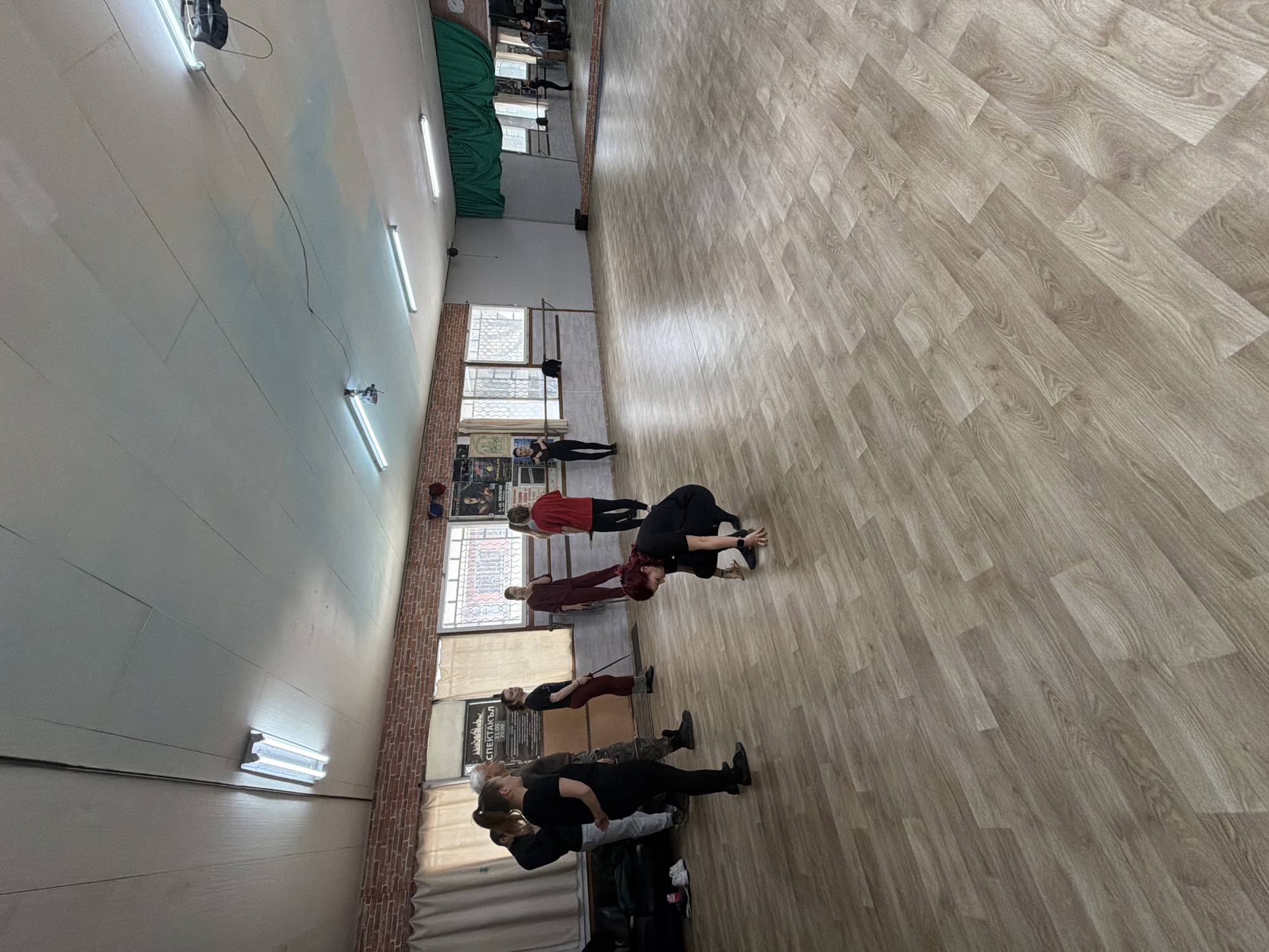 A group of people is engaged in a dance or exercise class in what appears to be a studio or dance hall. The room features a wooden floor, white walls, and fluorescent lighting. Along one wall, there are windows with posters, possibly advertisements for local events. The people are arranged in rows and performing synchronized movements. Based on visual landmarks and the apparent architecture in the background, this scene could be taking place in a studio located in Plovdiv, Bulgaria.