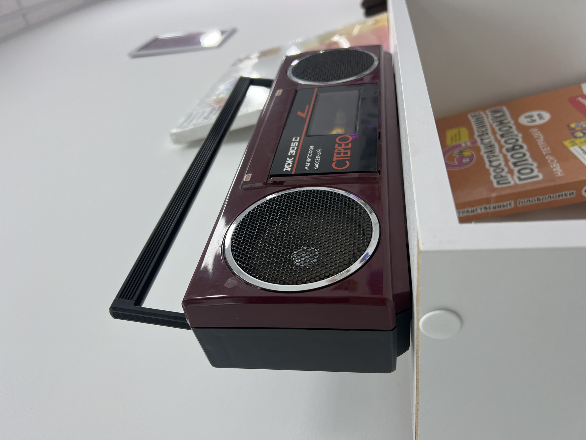 A vintage burgundy boombox sits on a white shelf, angled towards the viewer from a low, upward perspective. The boombox features two silver-rimmed speakers, a black cassette deck with "STEREO" printed in red, and a black retractable handle. To the right of the boombox, a blurred book with colorful illustrations and Cyrillic text, titled "Пространственные Головоломки" (Spatial Puzzles), is partially visible on the shelf below. The background consists of a plain white wall and shelf. The scene appears to be indoors, possibly in a home or retail setting. The image is sharp and well-lit, with the boombox being the clear focal point. The overall impression is one of nostalgia and retro technology.