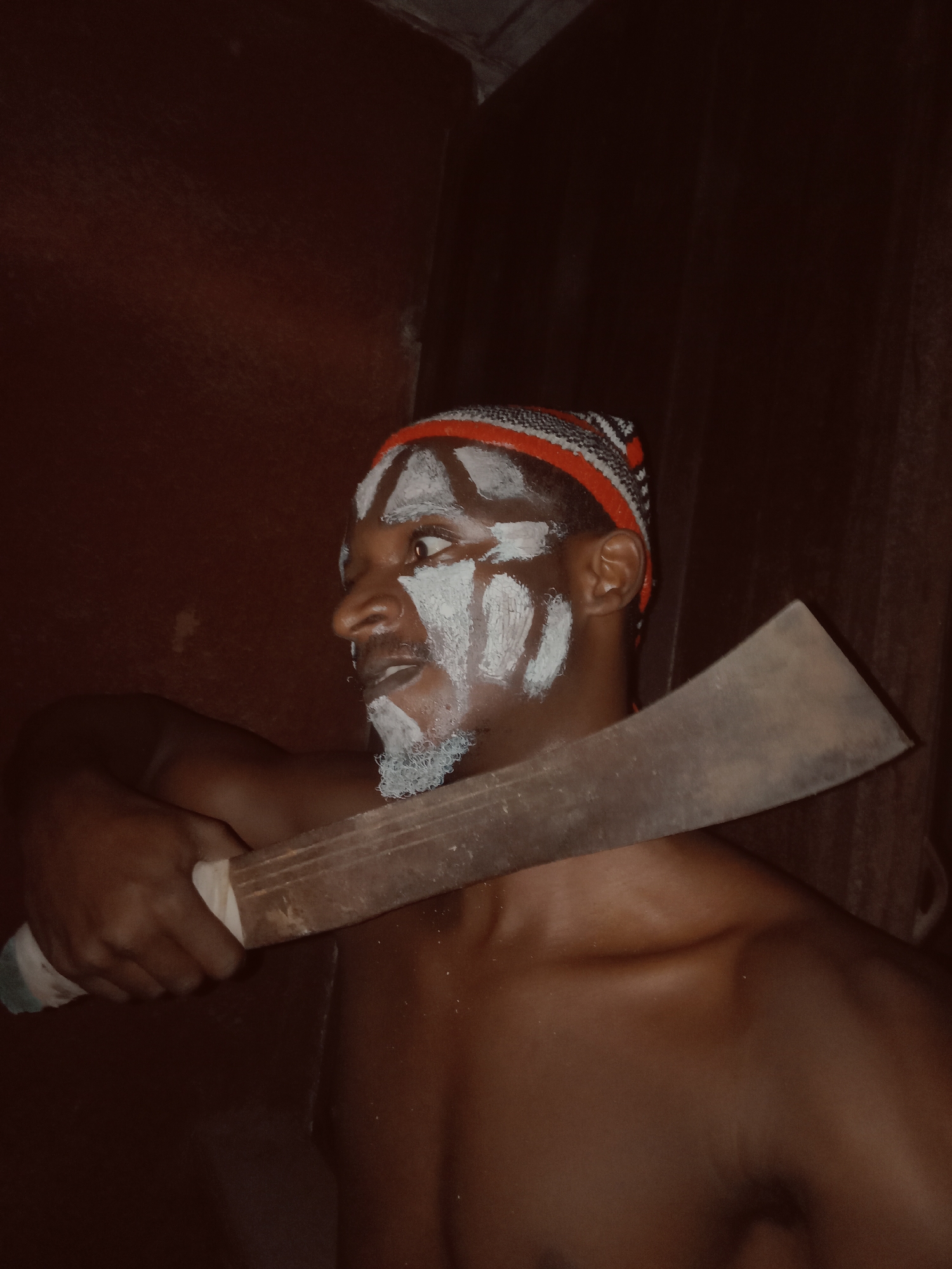 An individual, identified as a man, is depicted from the chest up in a dimly lit, enclosed space, possibly indoors or a confined area with dark brown walls. He is of dark complexion and is adorned with white face paint, applied in striking patterns across his forehead, cheeks, and chin, including a painted beard. He wears a knit head covering or cap with red, white, and black stripes.
The man holds a large, rustic machete or cutlass horizontally across his body, gripping the handle with his right hand which is wrapped in white material. His gaze is directed intensely towards the right side of the frame, giving a sense of alertness or focus. His bare torso is visible, showing well-defined shoulders and chest.
The scene suggests a cultural or ceremonial context, possibly a performance or ritual, given the traditional face painting and the presence of the machete, which can be a symbolic item in various African cultures. The lighting is low, creating a dramatic and somewhat mysterious atmosphere. No specific time of day or weather can be determined, but the indoor setting is implied. No text is visible in the image. The location context provided is Onitsha, Nigeria.