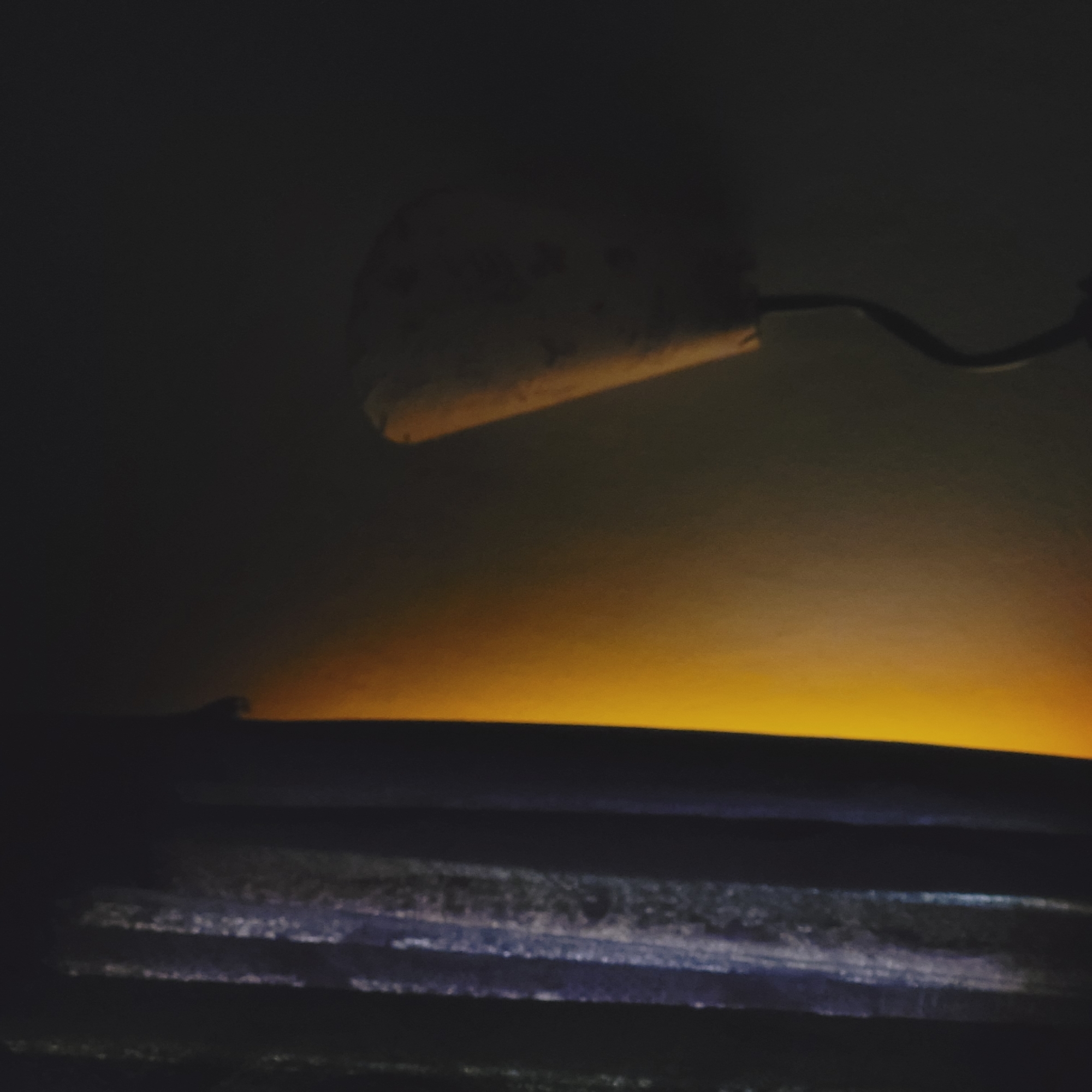 The image depicts a dark indoor scene, likely at night, characterized by a single prominent light source. In the upper left, a partially visible, light-colored lampshade or fixture emits a soft, warm yellow-orange glow. A dark, curved arm extends from this fixture, suggesting it's part of a lamp. This warm light illuminates a horizontal band across the middle of the frame, creating a striking gradient from bright yellow at its base to a softer orange as it fades into the dark background. Below this illuminated area, several dark, horizontal structures are visible, appearing as textured planks, shelves, or possibly blinds. These surfaces show subtle reflections of the yellow light and also exhibit faint blue-ish highlights. The overall atmosphere is quiet and intimate, with no discernible activity or visible text. The image does not contain specific visual cues to indicate its location in San Fernando del Valle de Catamarca, Argentina.