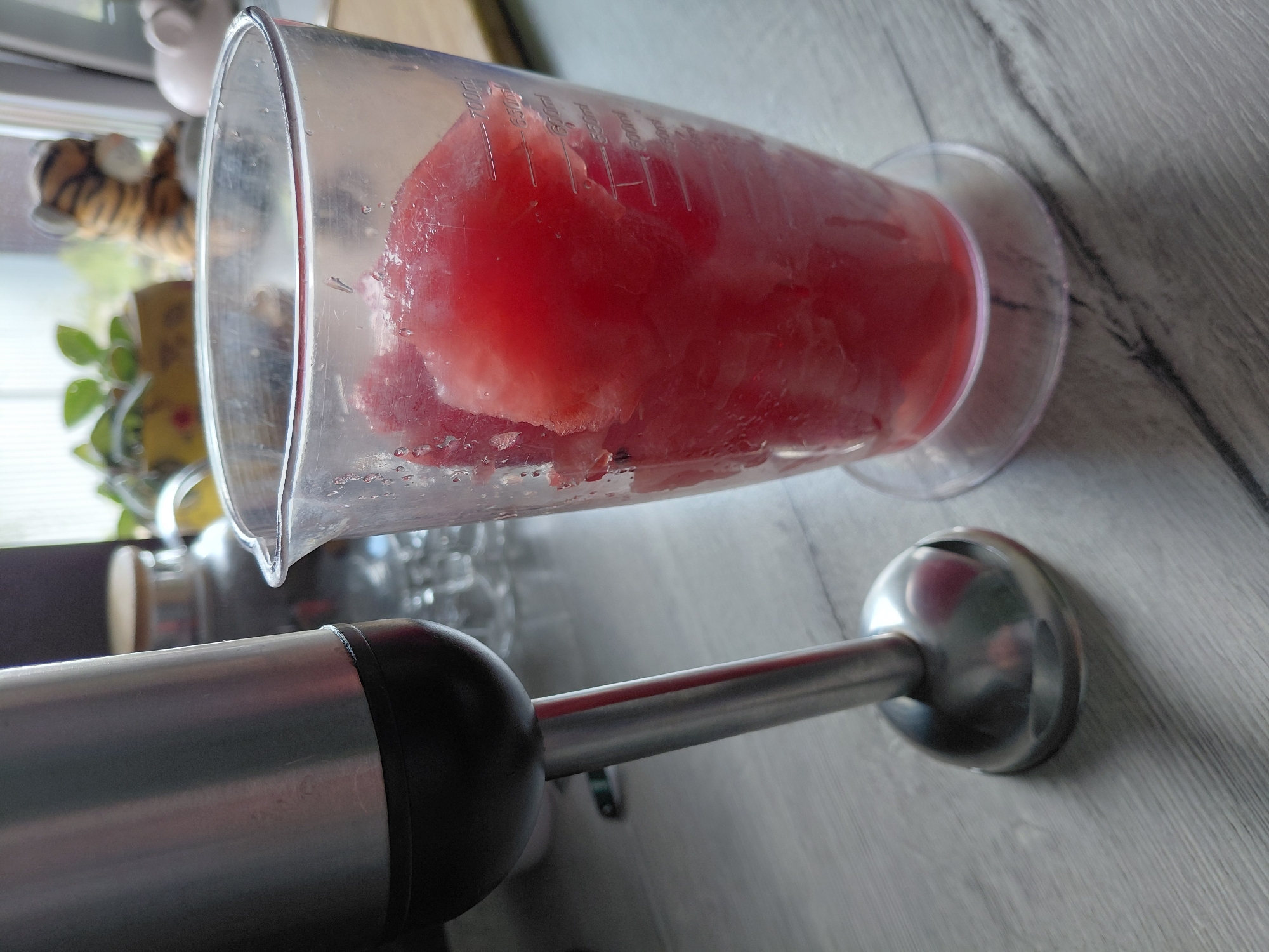 An immersion blender with a silver metal shaft and black grip rests on a grey wooden surface to the left of a clear plastic measuring cup. The measuring cup contains large chunks of red fruit, likely watermelon, and is marked with volume measurements up to 700 ml. In the background, a window shows a blurred outdoor scene with greenery. To the right of the window, a stuffed tiger toy and a pink pig figurine are visible. The scene appears to be indoors, possibly a kitchen. There are no people in the image. The overall impression is of food preparation.
