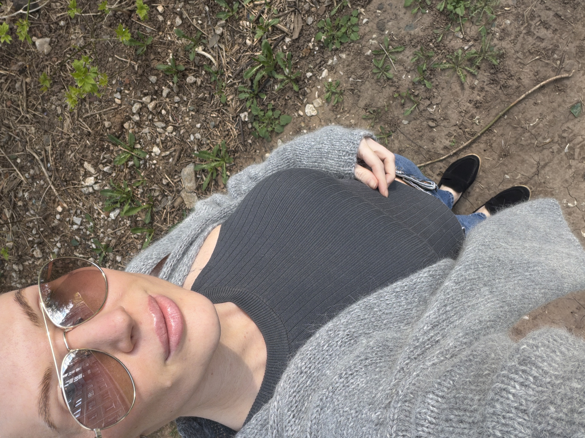 A top-down view of a person wearing sunglasses, a gray ribbed top, and a gray knit cardigan. The person is standing on bare earth scattered with small rocks and sparse vegetation. The person is wearing blue jeans and black loafers. The location is unspecified and cannot be determined to be Perm, Russia based solely on the image.