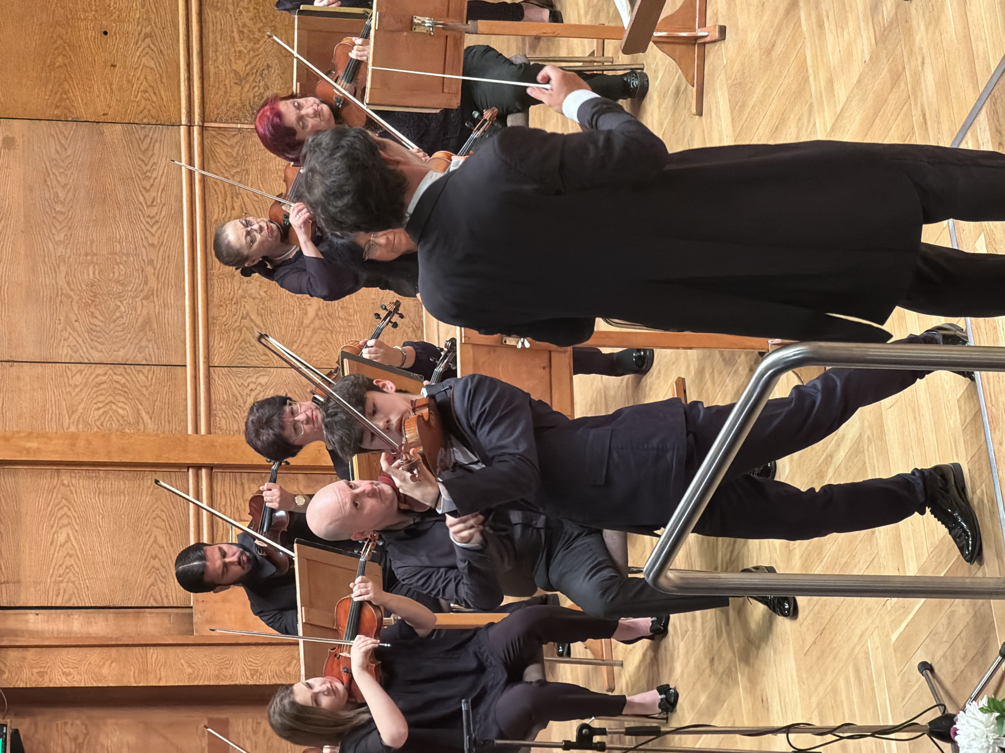 In a concert hall with wood-paneled walls, a youth in a dark suit stands in the foreground, playing a violin. He is positioned in front of other musicians who are also playing string instruments. A conductor, seen from behind in a black suit and white shirt, stands further back, holding a baton. The concert hall appears to be located in Sofia, Bulgaria. The lighting is bright, illuminating the musicians and the wooden stage.