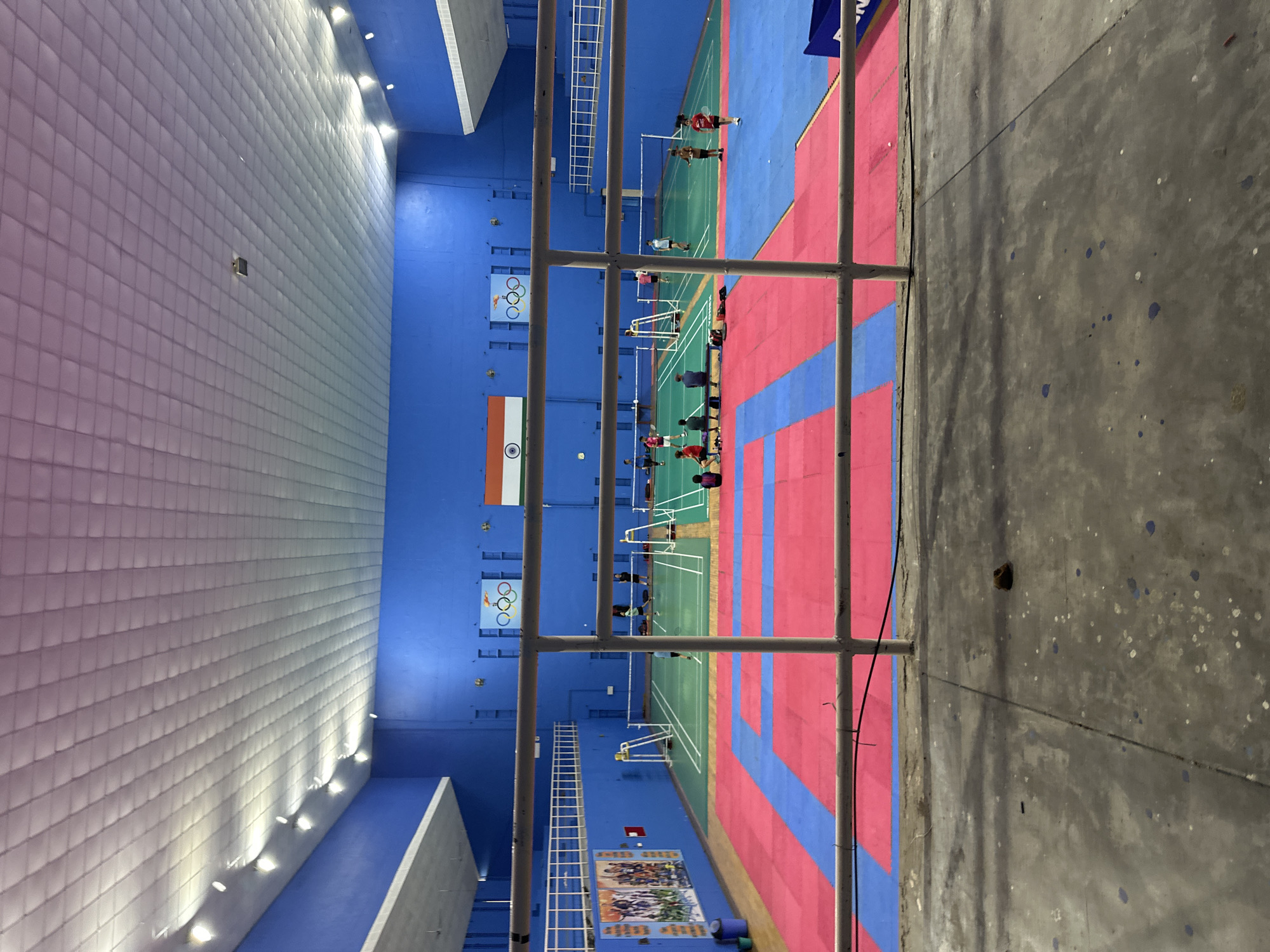 The image shows an indoor sports facility with multiple badminton courts and a taekwondo mat area. The walls are painted blue, and the ceiling is a white tiled structure with lights. The Indian flag and Olympic rings are visible on the wall. Several people are scattered across the courts, some playing badminton, others observing or resting. The taekwondo mat is divided into pink and blue sections. In the foreground, a metal railing runs horizontally across the frame, obscuring a portion of the scene. The floor in the foreground is concrete, showing signs of wear and some scattered debris.