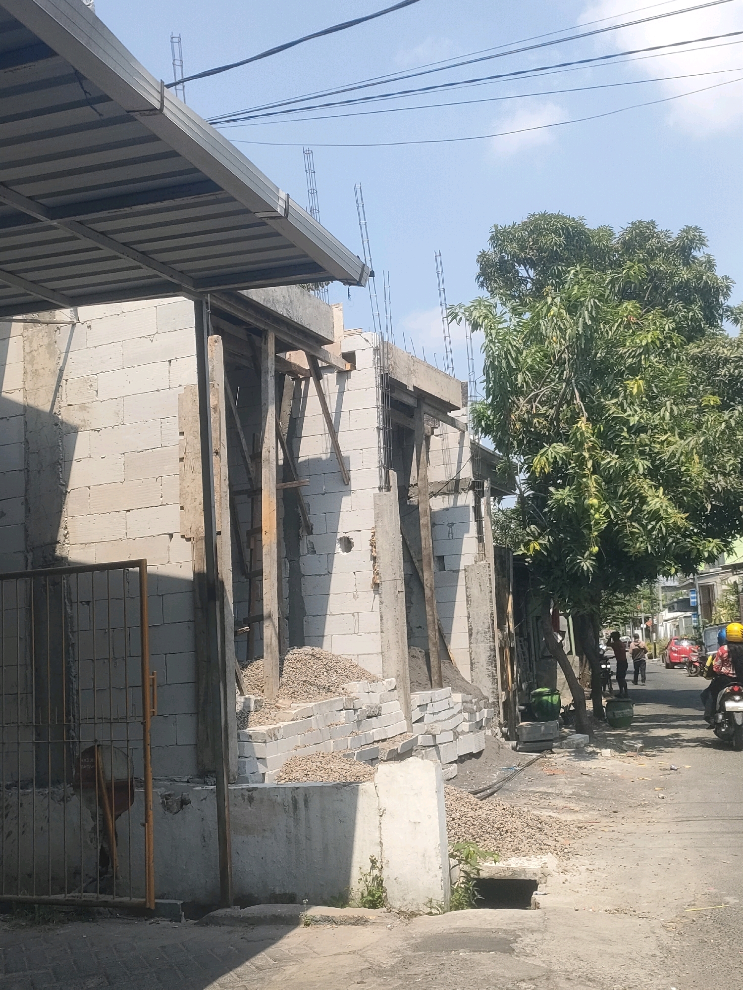 This is a medium-shot, eye-level, outdoor photograph taken on a clear day. The scene depicts a construction site on a street, likely in a residential or mixed-use area of Surabaya, Indonesia.
In the foreground and midground, a building is under construction. The walls are made of concrete blocks, and wooden scaffolding is in place. A corrugated metal roof structure is visible on the left side of the image, extending over the construction area. Piles of gravel and stacks of concrete blocks are scattered around the construction site, indicating ongoing work. A metal gate with a yellow frame is positioned on the far left, partially obscuring a concrete wall.
A large tree with dense green foliage stands to the right of the building, its branches reaching towards the construction. The street in the background is paved with asphalt and appears to be a relatively narrow road. There are several people on the street. Two individuals are standing near the tree, and a third person is walking away from the camera. On the right side of the street, a person is riding a motorcycle with a yellow helmet, and another motorcycle is parked behind them. A red car is also visible further down the street, parked near a building. Electrical wires crisscross the sky above the scene. The overall impression is one of an active urban construction environment.