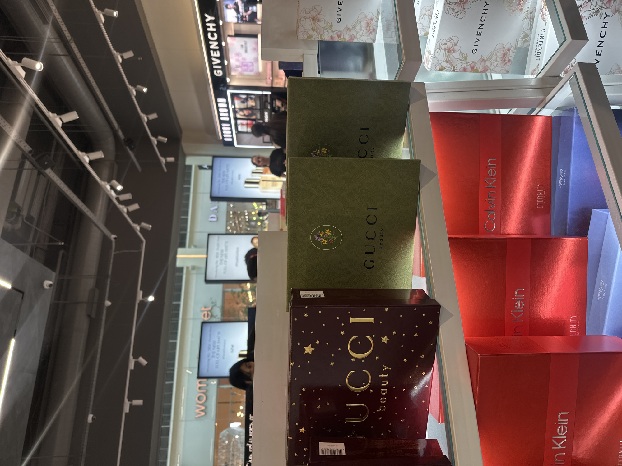 The image shows a display of perfume boxes inside a shop. The boxes are arranged on shelves. There are Gucci and Calvin Klein perfume boxes. In the background, there are displays of other brands like Givenchy and Bobbi Brown, as well as advertisements for "Wom" and "Da." The location is likely a duty-free shop or a department store selling cosmetics. There's a person browsing behind the perfumes. It is impossible to establish if the location is Yerevan, Armenia with only this visual information.
