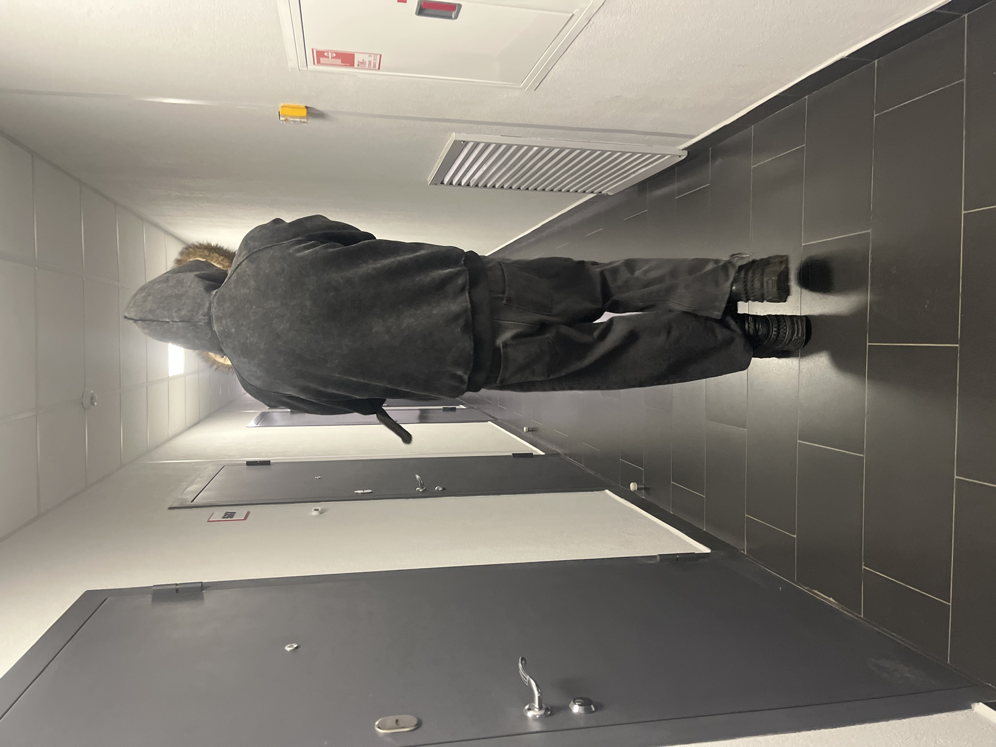 A person wearing a dark hooded jacket and dark pants is standing in a hallway with gray tiled floors and light gray walls. The person is facing away from the camera and toward the end of the hallway. There are several doors on the left side of the hallway, and a fire extinguisher box on the right side. The environment suggests an interior space, possibly a residential building or apartment complex in Saint Petersburg, Russia.
