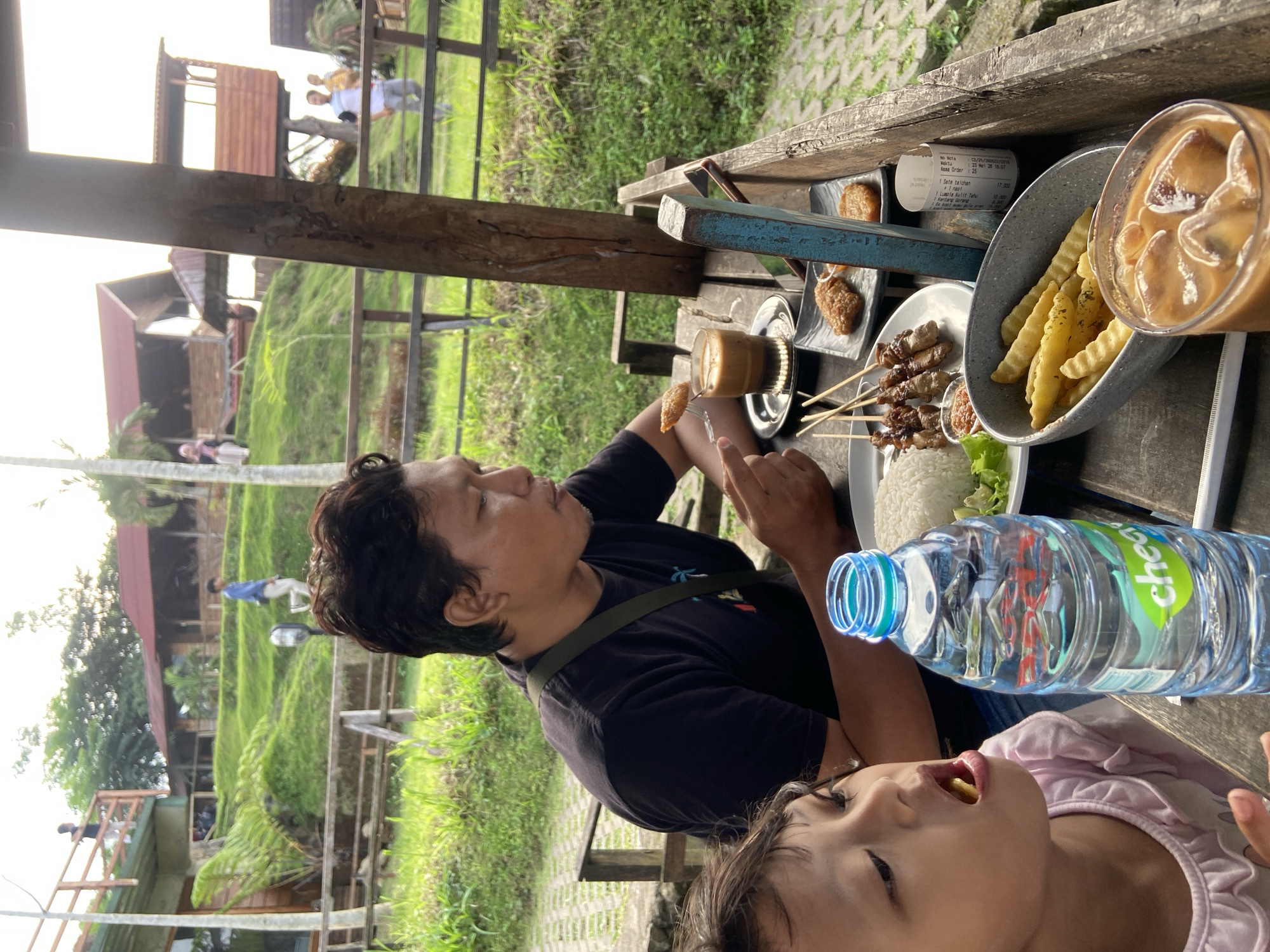 Here's a detailed description of the image:
The image shows a man and a child eating at a table in an outdoor setting. The man is holding a piece of food with a fork, and the child is eating a French fry. The table is covered with various dishes, including satay skewers, French fries, rice, and a glass of iced coffee. There is also a receipt and a bottle of water on the table. The background of the image shows a green, hilly landscape with buildings and other structures. The Nota indicates that this scene is occurring in Tamiajeng, Indonesia.