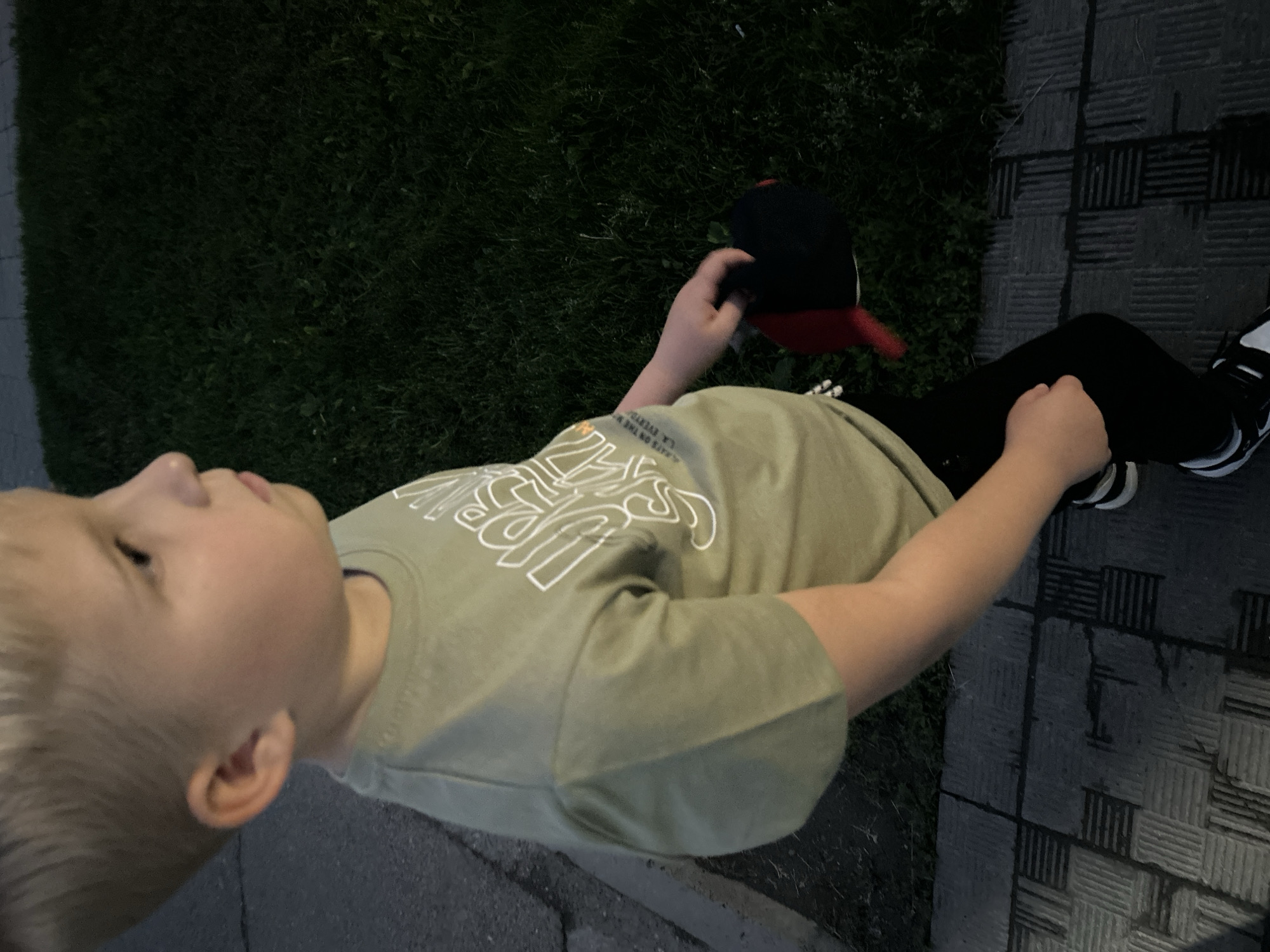 A young boy with blond hair stands outdoors, holding a red and black baseball cap in his right hand. He is wearing a light green t-shirt with white lettering and black pants. The boy is walking on a paved area next to a grassy patch. The scene appears to be at dusk, as the lighting is dim.