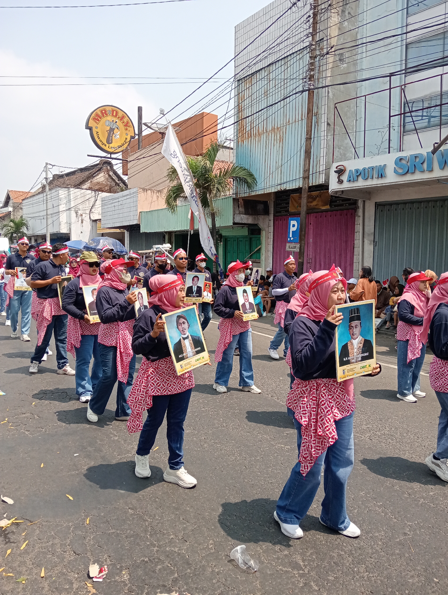 In the city of Demak, Indonesia, a parade is taking place on a sunny day on a paved street lined with buildings. A group of people, predominantly women wearing pink hijabs and red and white patterned sashes over navy blue shirts and jeans, are marching in formation. They are holding framed portraits of men. Several of the portraits are identifiable by text on the frames, such as "K.H. ABDURRAHMAN WAHID" and "DEPT. K." The parade appears to be organized and orderly, with participants walking in straight lines. In the background, several buildings are visible, including one with a prominent "APOTIK SRIWIJAYA" sign and another with a large, circular "MR. D.I.Y." sign. There are also bystanders watching the parade from the sidewalks. The overall atmosphere suggests a public event or celebration.