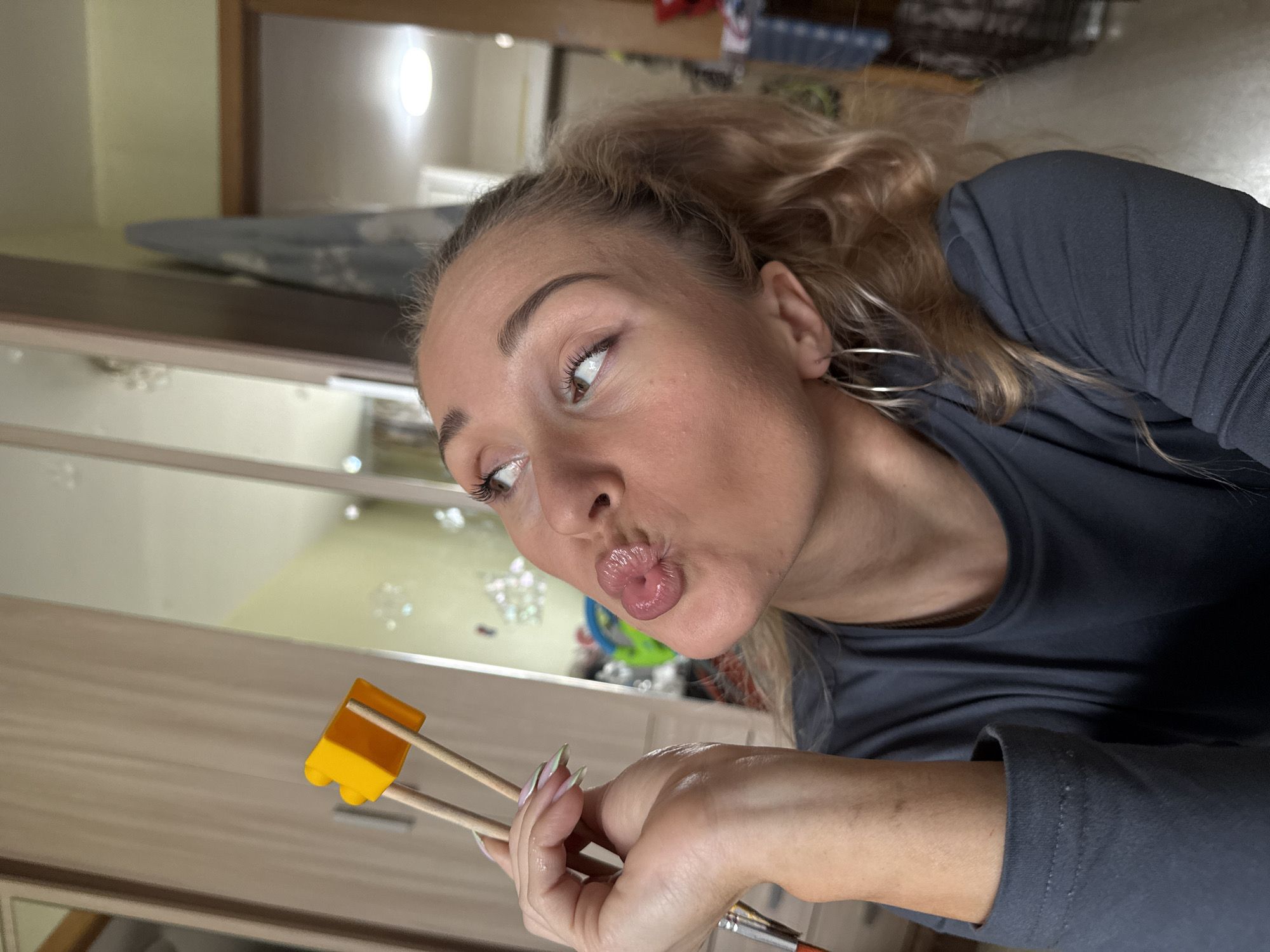 A young Caucasian woman with blonde hair in a ponytail is holding chopsticks with a small yellow object between them, near her face. She is puckering her lips and looking upwards. She is wearing a dark grey t-shirt and large hoop earrings. Her right hand, which is holding the chopsticks, has a paintbrush visible. The background appears to be an indoor setting with wooden cabinets and a mirror. The lighting is bright, suggesting daytime. The location is not discernible from the image.