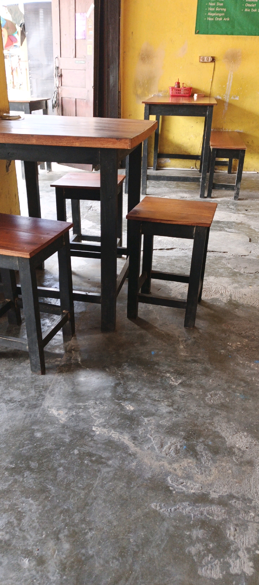 The image shows a simple outdoor seating area at a food stall or small restaurant in Godean, Indonesia. The scene is captured from a low angle, focusing on two tables and their accompanying stools. The tables are made of dark wood with black legs, and the stools have wooden seats and dark legs.
The foreground features a dark wooden table with two stools. One stool is directly in front of the table, and another is partially visible to its left. The floor is a rough, textured concrete surface with visible wear and tear.
In the background, a second, smaller table stands against a bright yellow wall. This table also has dark legs and a wooden top, and a red basket containing two bottles of sauce is placed on it. A single stool is visible next to this table. The yellow wall has some light-colored marks and what appears to be a faint water stain. Above the yellow wall, a green sign with text is partially visible, likely indicating the menu.
To the left of the scene, a wooden door with vertical planks is visible, with some items hanging on it. Beyond the door, the interior of a building is hinted at, with glimpses of light and what might be decorative items. The overall impression is of a humble, functional dining space.