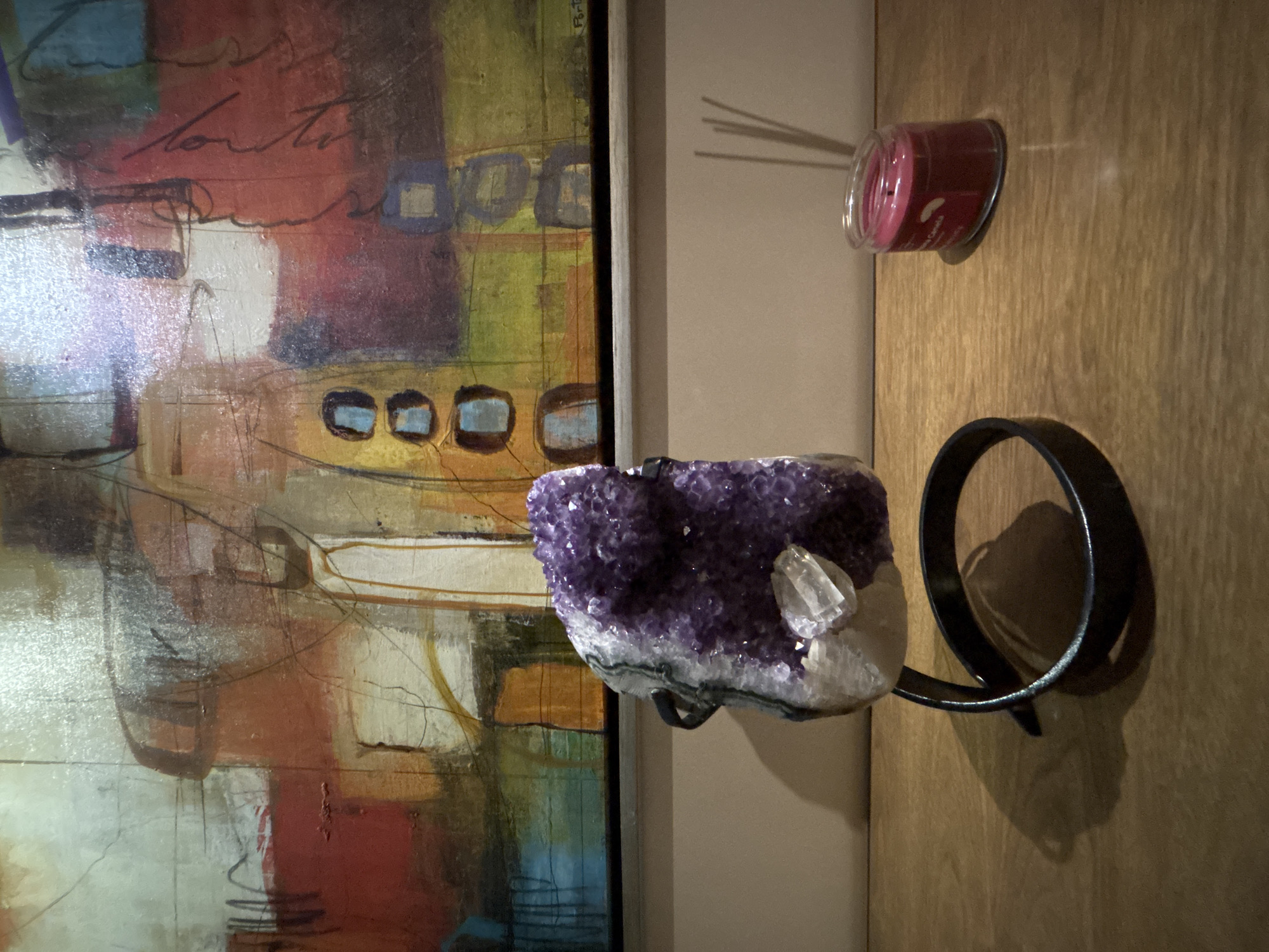 The image shows a section of a room interior. On the left is a framed abstract painting with various colors including red, white, blue, and yellow. Beside the painting is a wall upon which sits a black metal circular holder with a geode crystal cluster in the center. The crystal is primarily purple with a patch of white. Above it is a candle jar, filled with a pink substance and placed on a wall-mounted shelf. The wall is a light tan wood grain. The photo does not suggest any information about San Bernardino Tlaxcalancingo, Mexico.