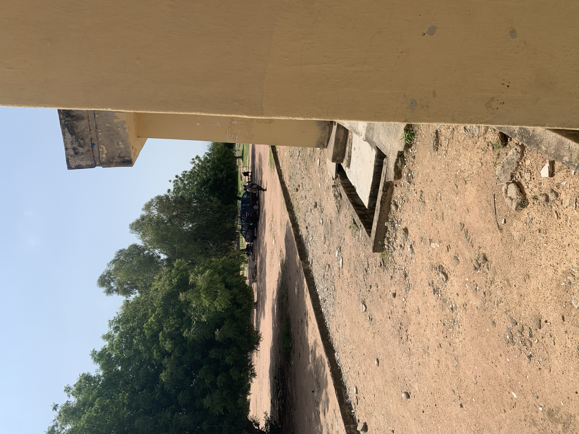The image depicts an outdoor scene in Jalingo, Nigeria, on a clear, sunny day. In the foreground, a portion of a yellow-painted building is visible on the right, its textured wall showing some wear and tear. Adjacent to the building, a concrete drainage system with several steps leads down to a recessed channel. The ground is covered in reddish-brown soil with scattered stones and some sparse vegetation. A low concrete curb lines a dirt path that extends into the midground. Beyond this path, several large trees with dense green foliage provide shade. In the background, there are a few dark-colored vehicles parked on the dirt ground, and two individuals can be seen standing near them. The overall impression is that of a utilitarian outdoor area, possibly on the grounds of an institution or public space, with a dry, dusty environment characteristic of the region.