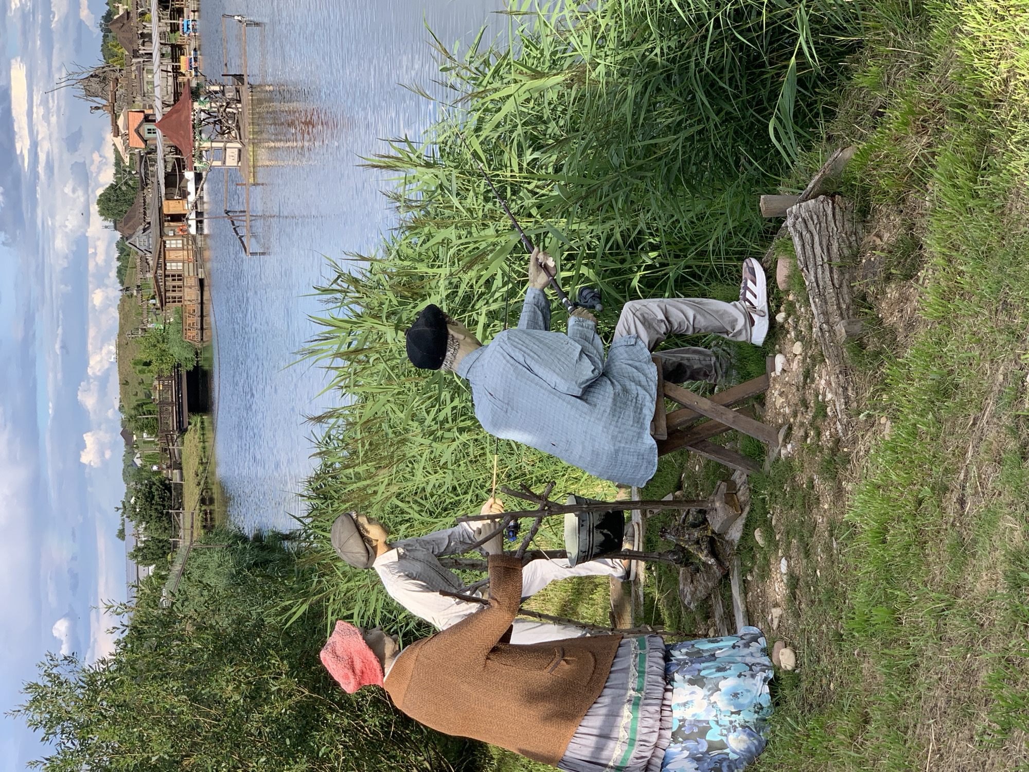 In the city of Malakhovetski Sel'ski Sovet, Belarus, an outdoor scene depicts three mannequins engaged in fishing by a lake. The mannequin on the left is dressed in a brown knitted cardigan, a long blue and white floral skirt, and a pink knitted hat, and is standing with its hands resting on a wooden support. Next to it, a mannequin in a light-colored long-sleeved shirt and cap is holding a fishing rod. The third mannequin, seated on a wooden stool and wearing a blue collared shirt, gray trousers, and a black cap, is actively fishing with a rod and reel. The mannequins are positioned amidst tall green reeds and grass. The lake extends into the background, where a picturesque village with wooden houses and a bridge is visible across the water. The sky is partly cloudy, with fluffy white clouds against a pale blue backdrop. The overall atmosphere suggests a tranquil, rural setting.