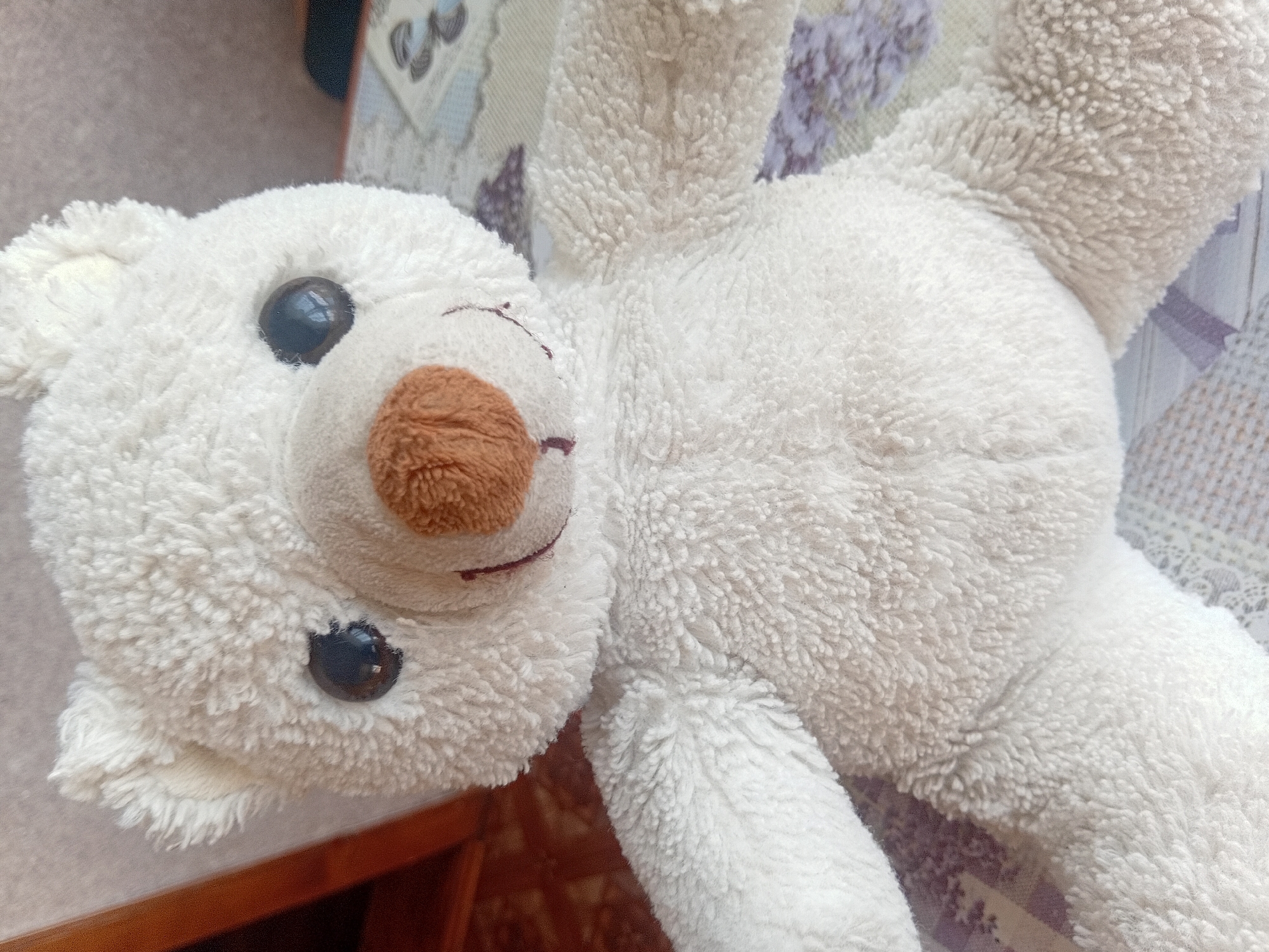 The image shows a close-up, high-angle view of a white, fluffy teddy bear lying on its back. The bear's face is to the left, with two black button eyes and a brown, heart-shaped nose. A small, stitched red mouth is visible below the nose. The bear's fur is soft and textured, with visible stitching details on its ears and paws.
The bear is lying on a surface that appears to be a patterned fabric, possibly a couch or blanket, with a floral or abstract design in shades of purple and beige. A wooden surface is visible in the lower left corner, suggesting the bear is on furniture. The background is softly blurred, with hints of what might be other fabrics or objects. The lighting suggests it's daytime, likely indoors. No text is visible in the image. The overall impression is of a soft, well-loved toy.