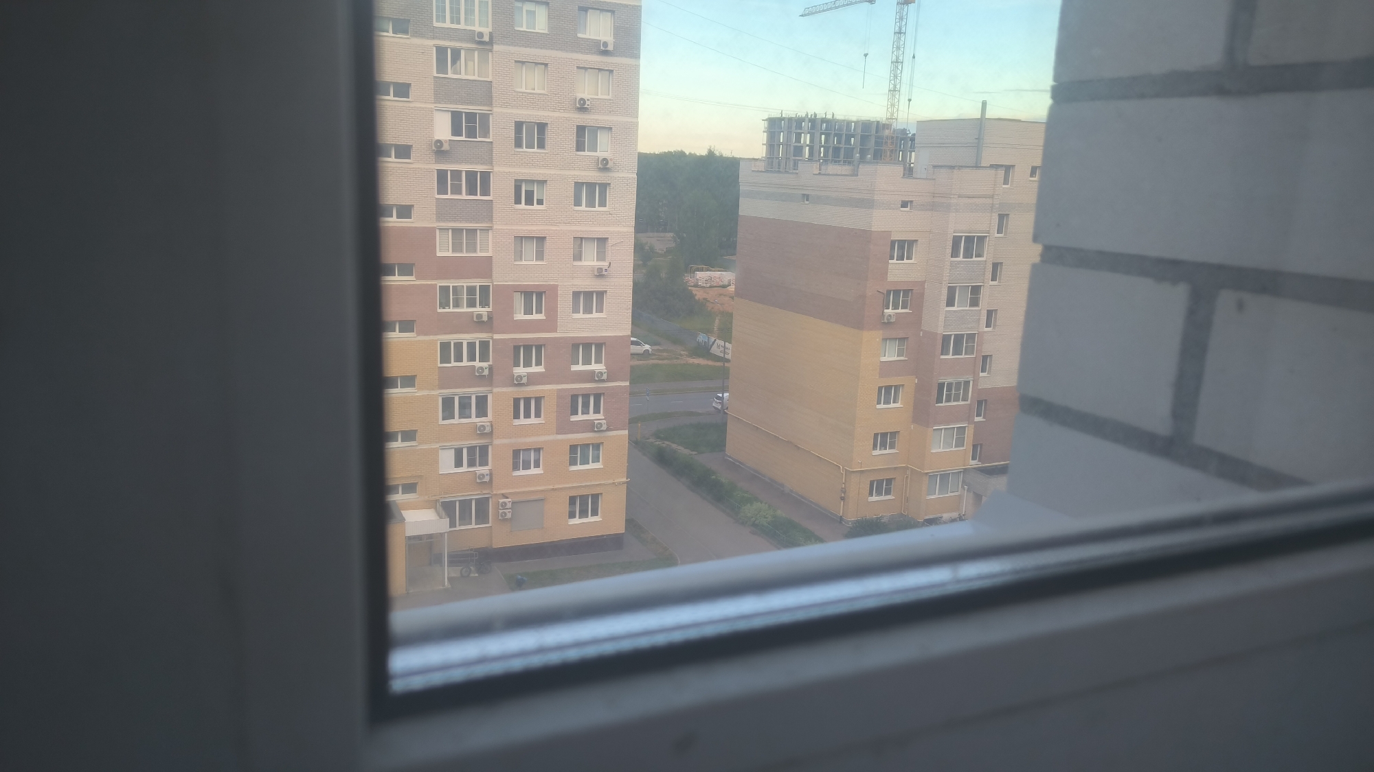 This is a medium-distance, eye-level exterior shot taken through a window, looking out at two tall, multi-story residential buildings in the city of Kovrov, Russia. The building on the left is light beige and white, with rows of windows and visible air conditioning units. The building on the right is a warm beige and brown color, also featuring numerous windows. Both buildings appear to be apartment complexes. In the background, there is a construction site with a crane and partially built structures, suggesting ongoing development in the area. Trees and a road with a few cars are also visible, indicating a residential neighborhood. The sky is a pale blue with a hint of haziness, suggesting it might be early morning or late afternoon. The foreground shows a portion of a white window frame, slightly out of focus.