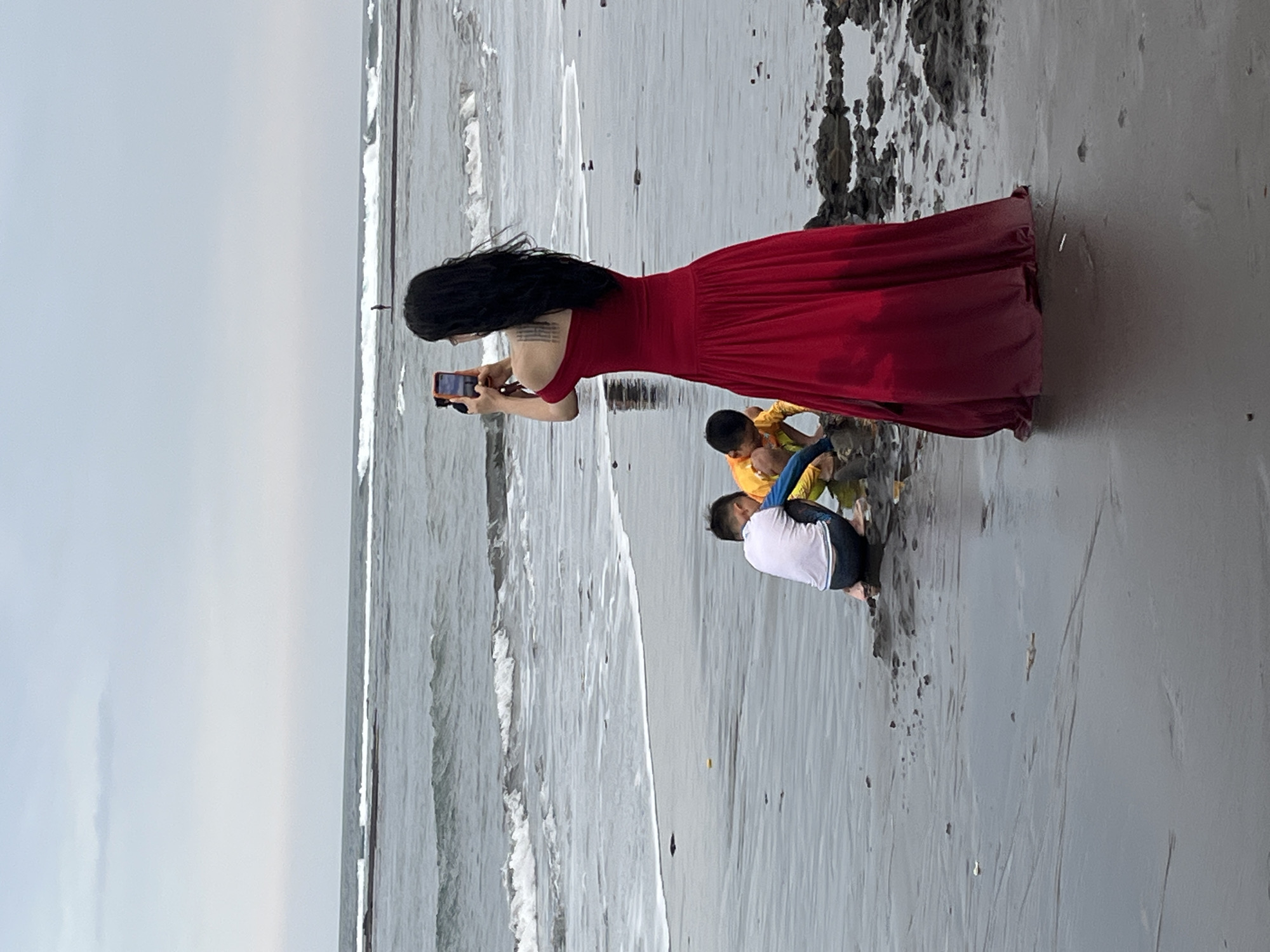 The image shows a woman in a long, red, off-the-shoulder dress standing on a beach and taking a photo with her phone. Two young boys are playing in the sand in front of her, closer to the water's edge. The beach is wet sand with some dark, rocky debris. The ocean is visible in the background with moderate waves and a cloudy sky. The scene is located on the beach in Blahbatuh, Indonesia.