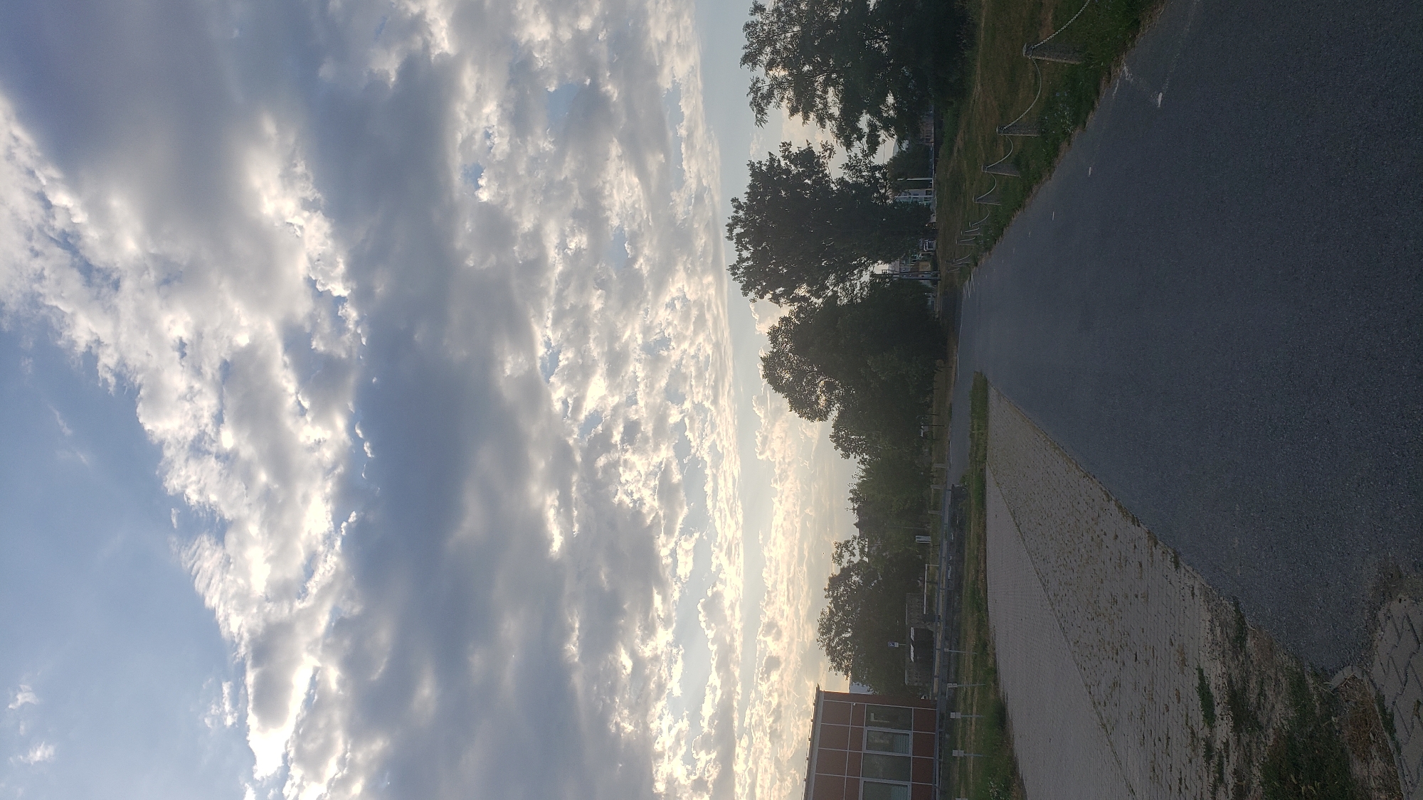 The image depicts an asphalt road leading into the distance, bordered by a grassy area with a rope fence on the right and a paved area with some vegetation on the left. A red-sided building is visible on the far left. Several trees are scattered throughout the scene, with a dense line of trees in the middle ground and more scattered trees on the right. The sky is filled with dramatic clouds, with patches of blue sky visible and the sun appearing to be setting or rising, casting a warm light through the clouds. The scene appears to be an outdoor pathway or road within an urban or semi-urban environment. No people or animals are visible in the image. The location is not identifiable as Prešov, Slovakia, without further information.