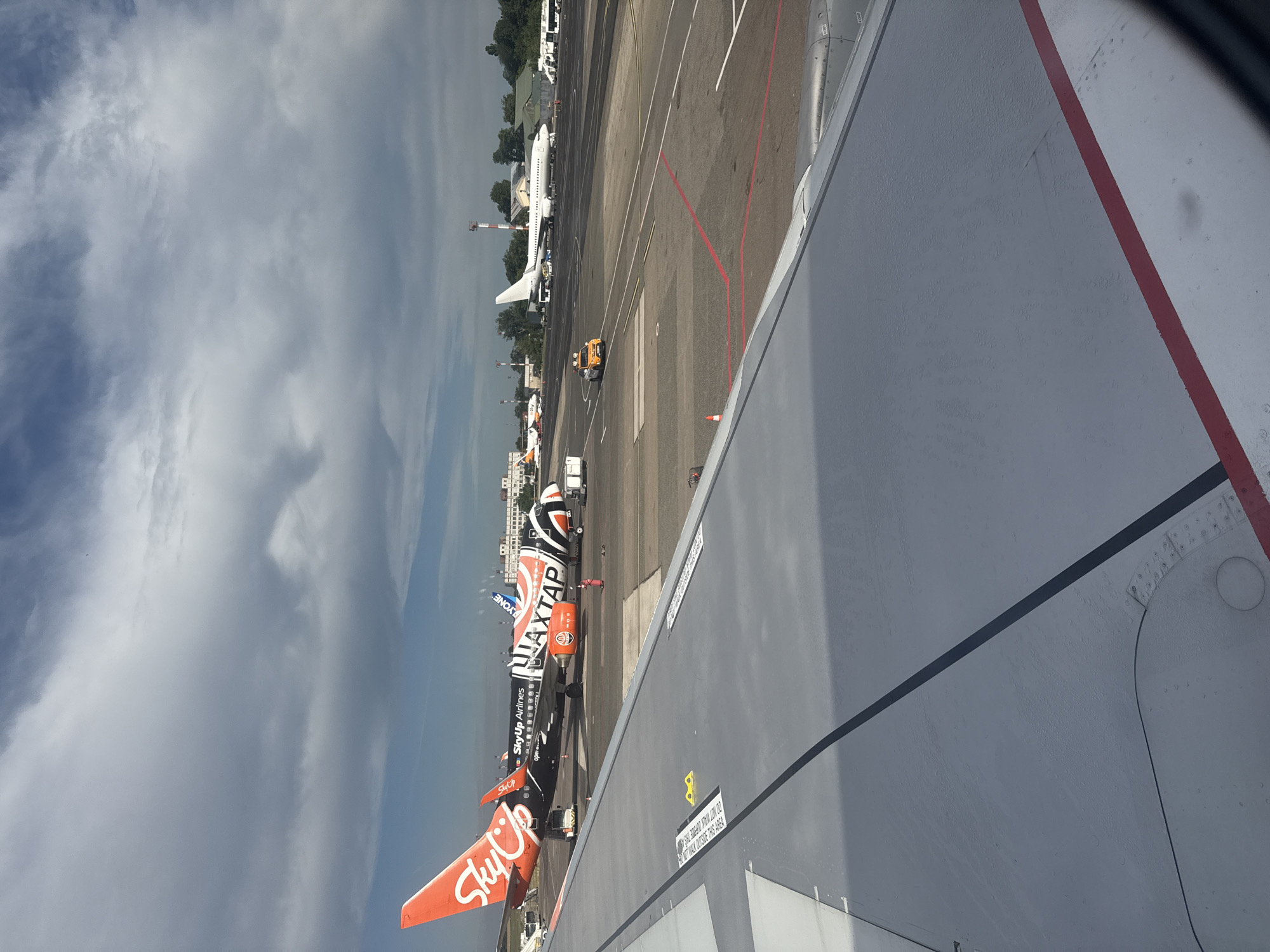 An aerial view from an airplane window shows a tarmac at the Chișinău International Airport. In the foreground, a portion of the airplane wing is visible, with a red and white stripe along its edge. Several airplanes are parked on the tarmac. One prominent airplane in the middle ground is black with orange, white, and striped orange and white accents. It displays "SkyUp Airlines" branding and "UAXTAP" in large orange letters. Another smaller airplane with orange and white livery is visible to the left. Further in the background, a white airplane is parked. Several airport vehicles, including a small yellow tug, are on the tarmac. The scene is set under a cloudy sky with patches of blue.