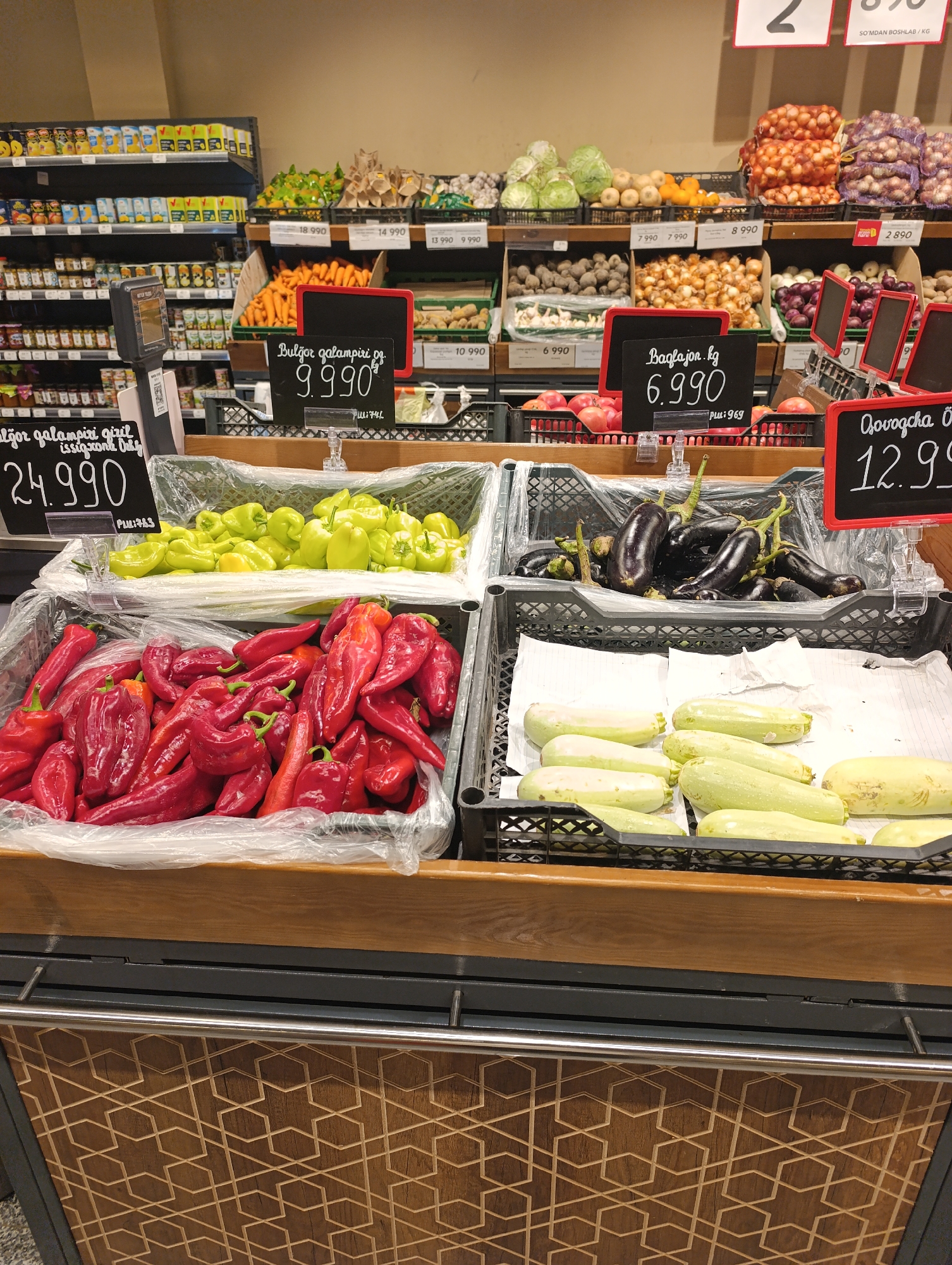 The image displays a produce section of a supermarket in Jizzakh, Uzbekistan. The foreground features several plastic crates filled with fresh vegetables. From left to right, the crates contain bright red peppers, followed by a crate of light green peppers. To the right, a crate holds dark purple eggplants, and beneath them, another crate is filled with pale green zucchini and a larger, light yellow squash. Price tags are visible for some of the produce, including "Bulgor qalampiri" priced at 9.990 and "Baglajon" at 6.990. In the background, shelves are stocked with various food items, including canned goods, jars, and what appear to be bags of onions and potatoes. The overall environment suggests a well-organized retail setting.