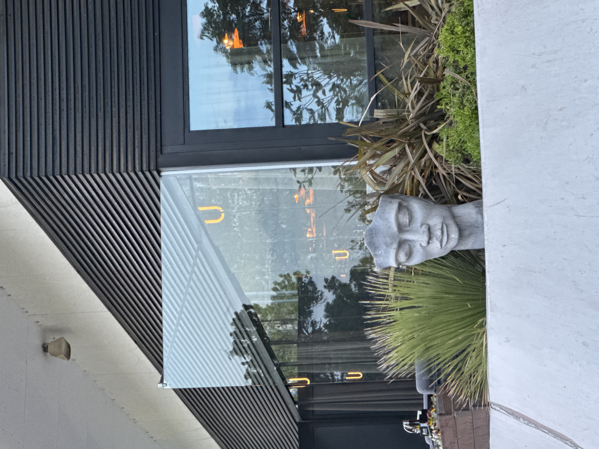 The image shows the exterior of a building in Burgas, Bulgaria. The building has a modern design with dark grey wooden slats on the facade, large windows with reflections, and a white concrete wall on the right side of the frame. A stone head statue is positioned next to the wall, surrounded by foliage. The reflection in the windows indicates an urban environment with trees and street lighting.