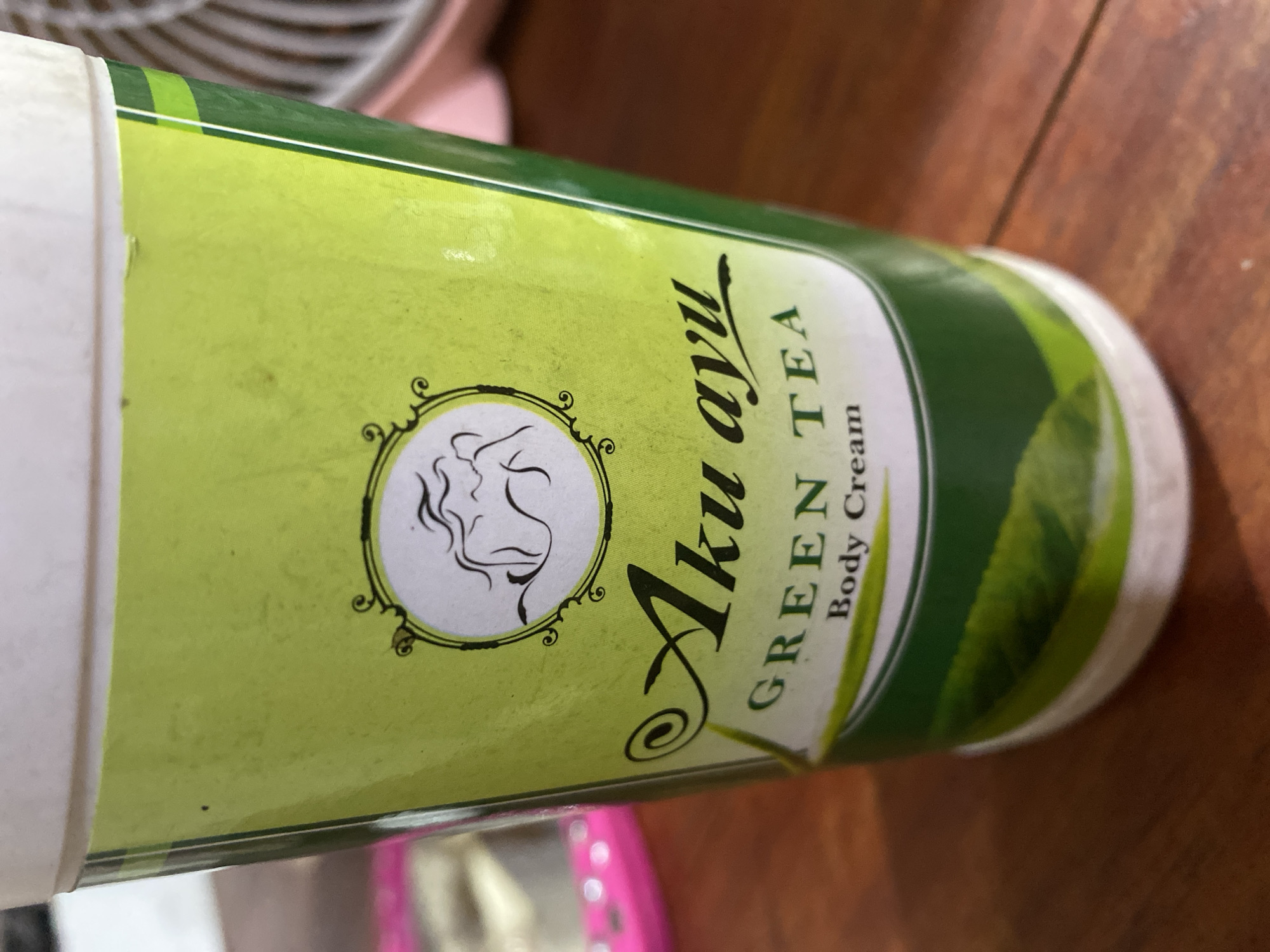The image shows a close-up, eye-level shot of a green tube of "Aku Ayu GREEN TEA Body Cream". The tube has a white cap and is set against a wooden surface. The branding is prominent, with the words "Aku Ayu" in a stylized black font, followed by "GREEN TEA" and "Body Cream" in smaller text. A circular emblem with a black outline contains a stylized depiction of a human figure. The background is blurred, showing hints of other objects, including what appears to be a pink item on the left and a white fan on the right, suggesting an indoor setting. The lighting is moderate, with some shadows present. The overall impression is that of a product shot, focusing on the packaging of the body cream. The location in Ta', Indonesia, is not visually discernible from the image itself.
