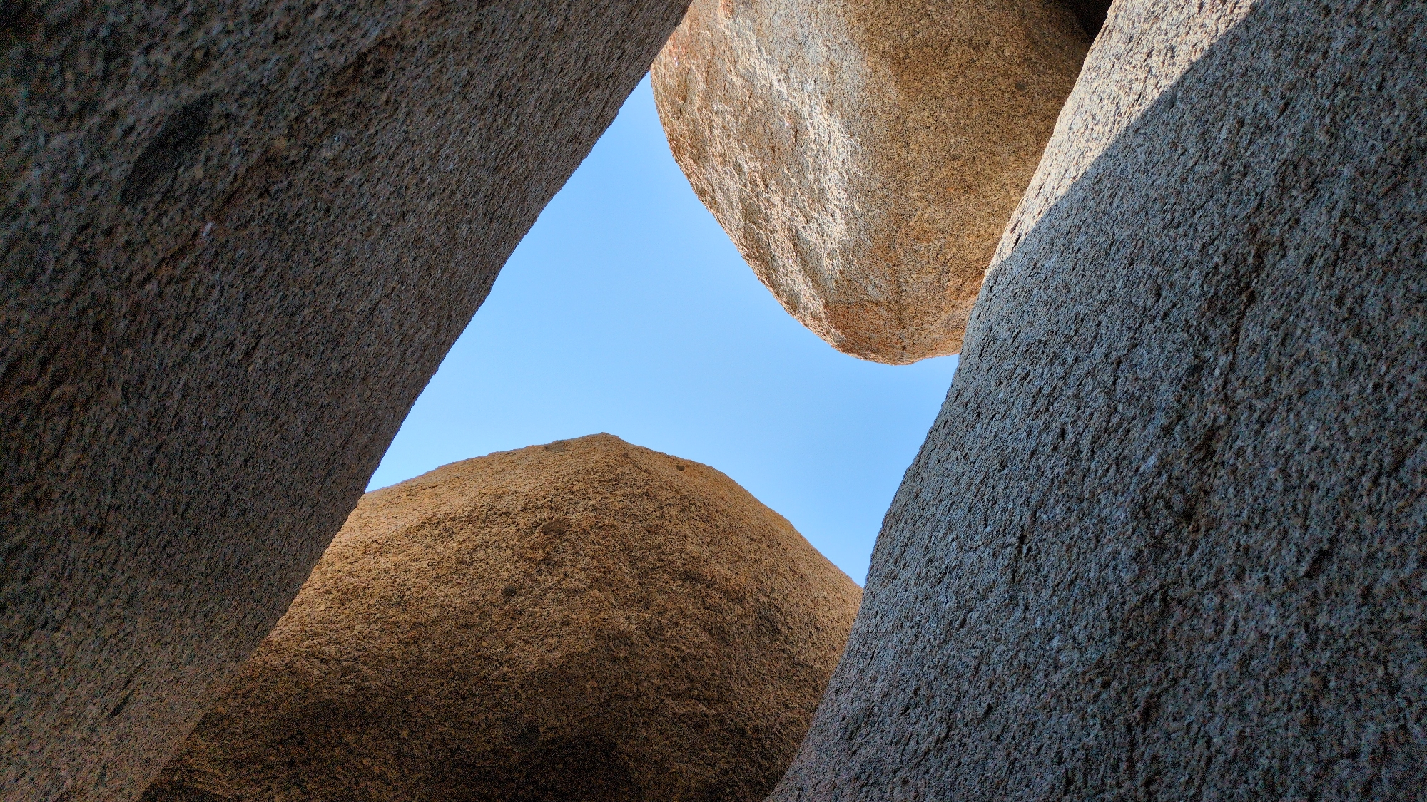 The image captures a ground-level view looking upwards through a narrow opening formed by large, textured rock formations. The rocks appear to be natural, with a rough, granular surface. A patch of clear, blue sky is visible through the opening. The color of the rocks varies from a darker, gray-brown to a lighter, tan shade. The scene evokes a sense of being enclosed within a rocky landscape. The location is Phuoc Dinh, Vietnam.