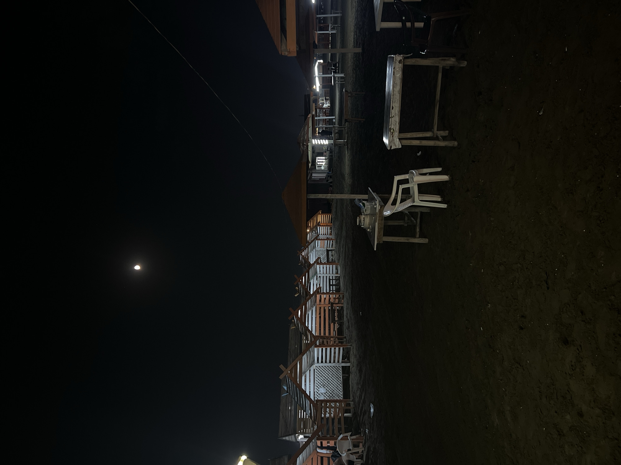 The image depicts a nighttime scene on a beach in Khazar Raion, Azerbaijan. Several small, open-sided wooden huts with sloped roofs line the sand, appearing to be part of a beachside establishment. In front of these huts, several tables and chairs are set up. Some tables are covered with white cloths, and some chairs are made of plastic. In the background, larger, covered seating areas with umbrellas are visible, suggesting a dining or relaxation area. A bright moon is visible in the dark night sky, and a thin wire stretches across the upper right portion of the frame. The ground is covered in sand. There are no people clearly visible in the scene.